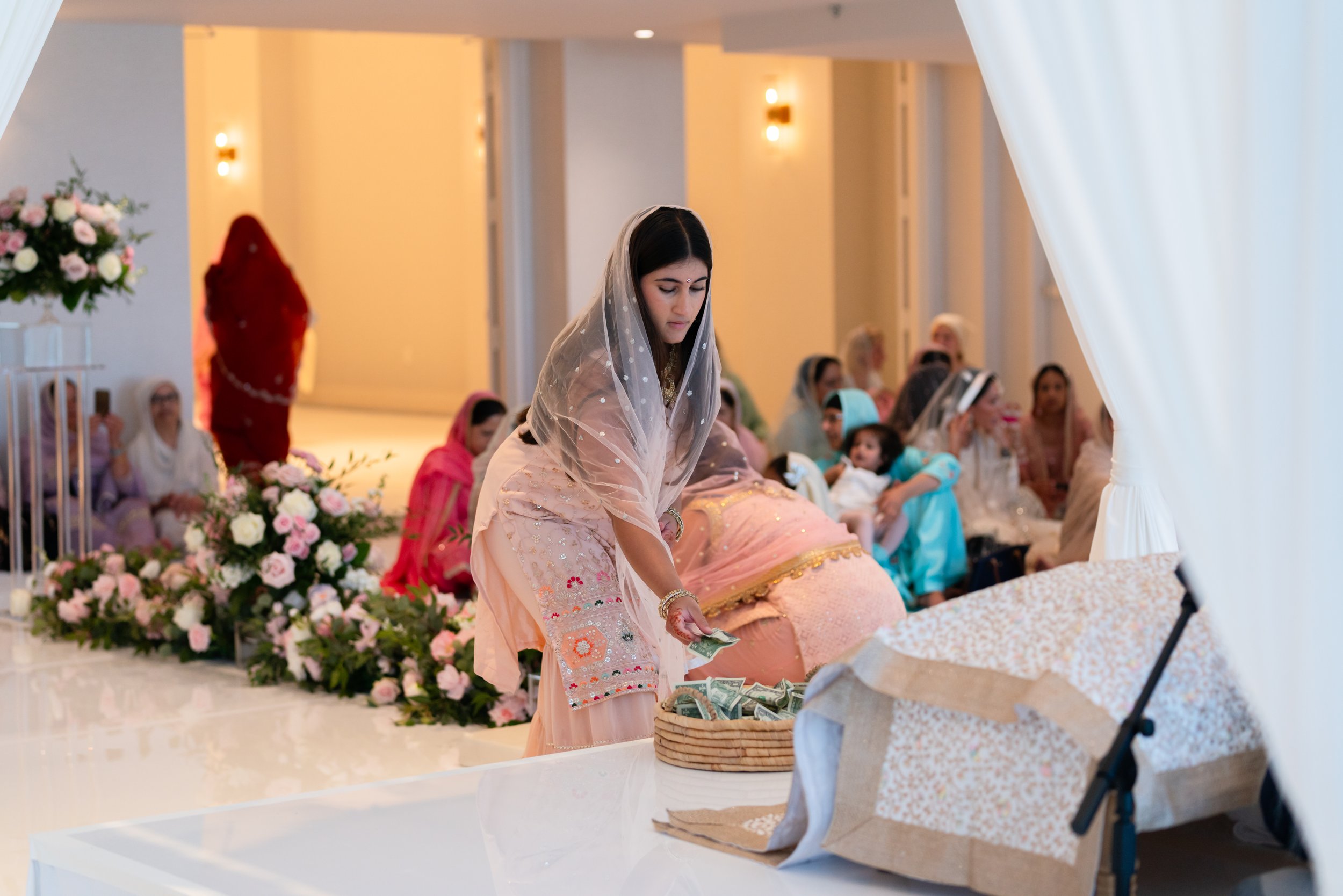A woman in traditional South Asian attire placing money into a basket at a religious or cultural ceremony with people sitting on the floor in the background.