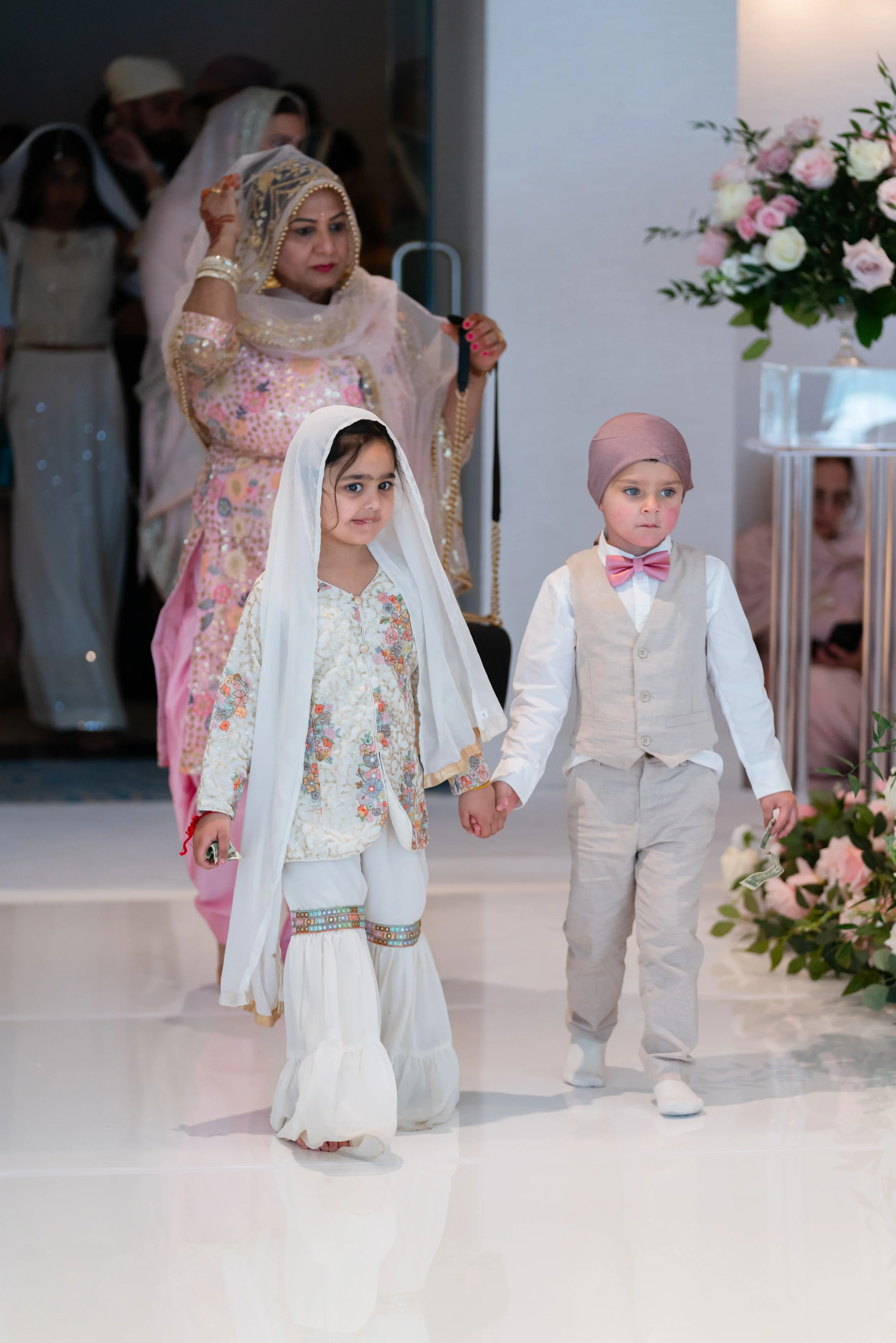 Young girl and boy dressed in traditional South Asian attire holding hands at a wedding or cultural event, with women in colorful outfits and floral decorations in the background.