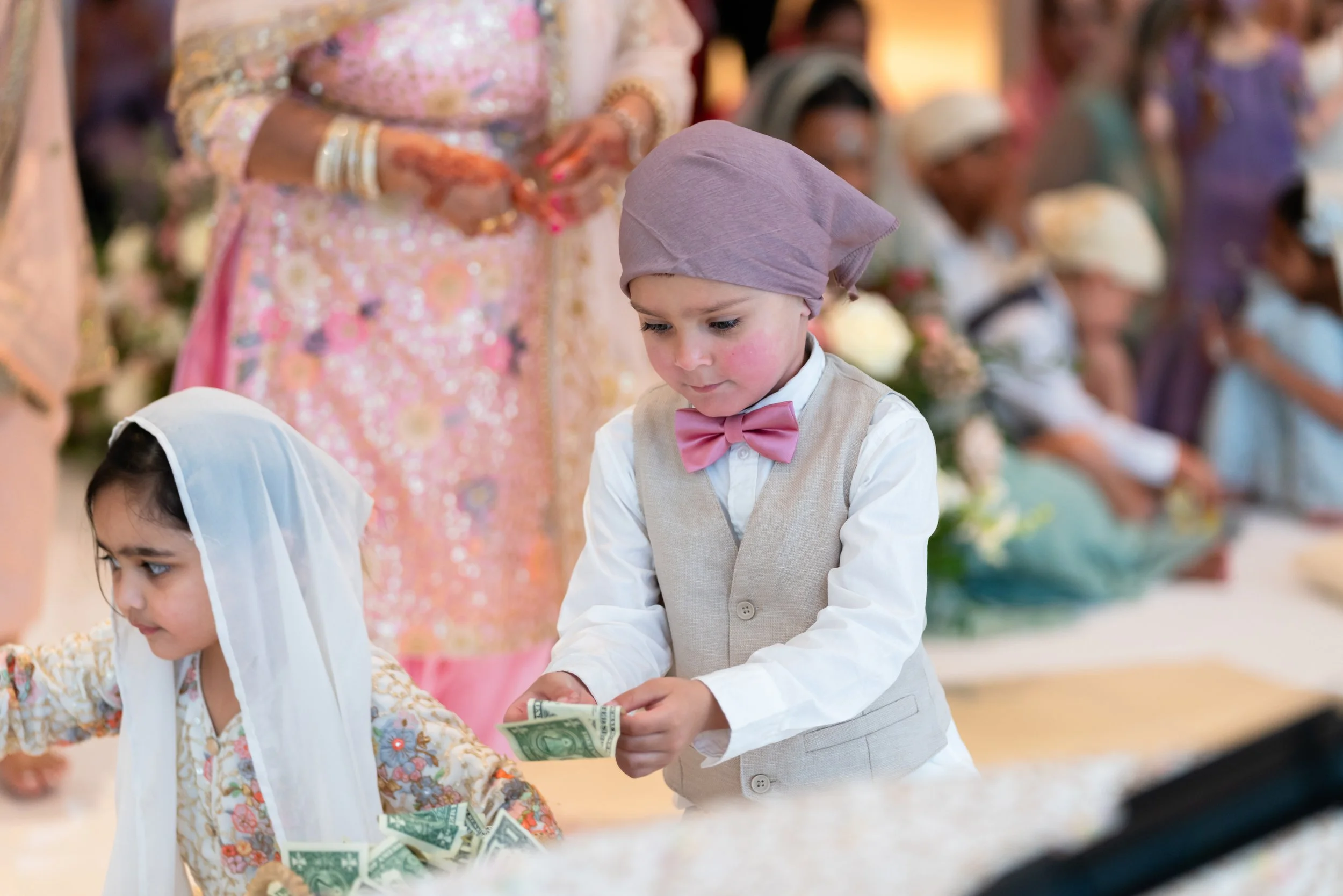 Young children dressed in traditional attire exchanging money at a cultural or religious ceremony, with adults and others in similar attire in the background.