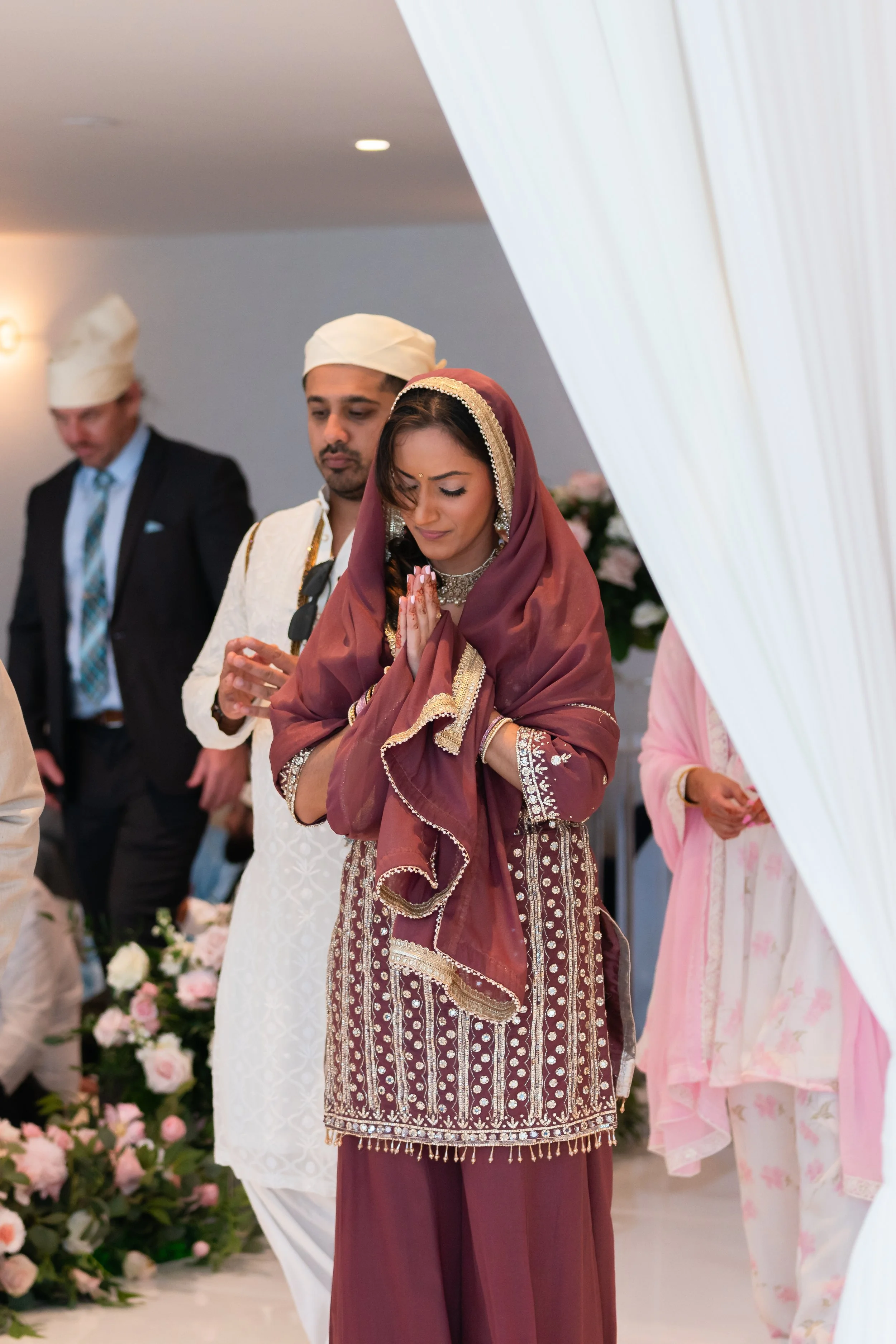 A woman dressed in traditional South Asian attire praying with her hands folded during a wedding ceremony, surrounded by other guests.