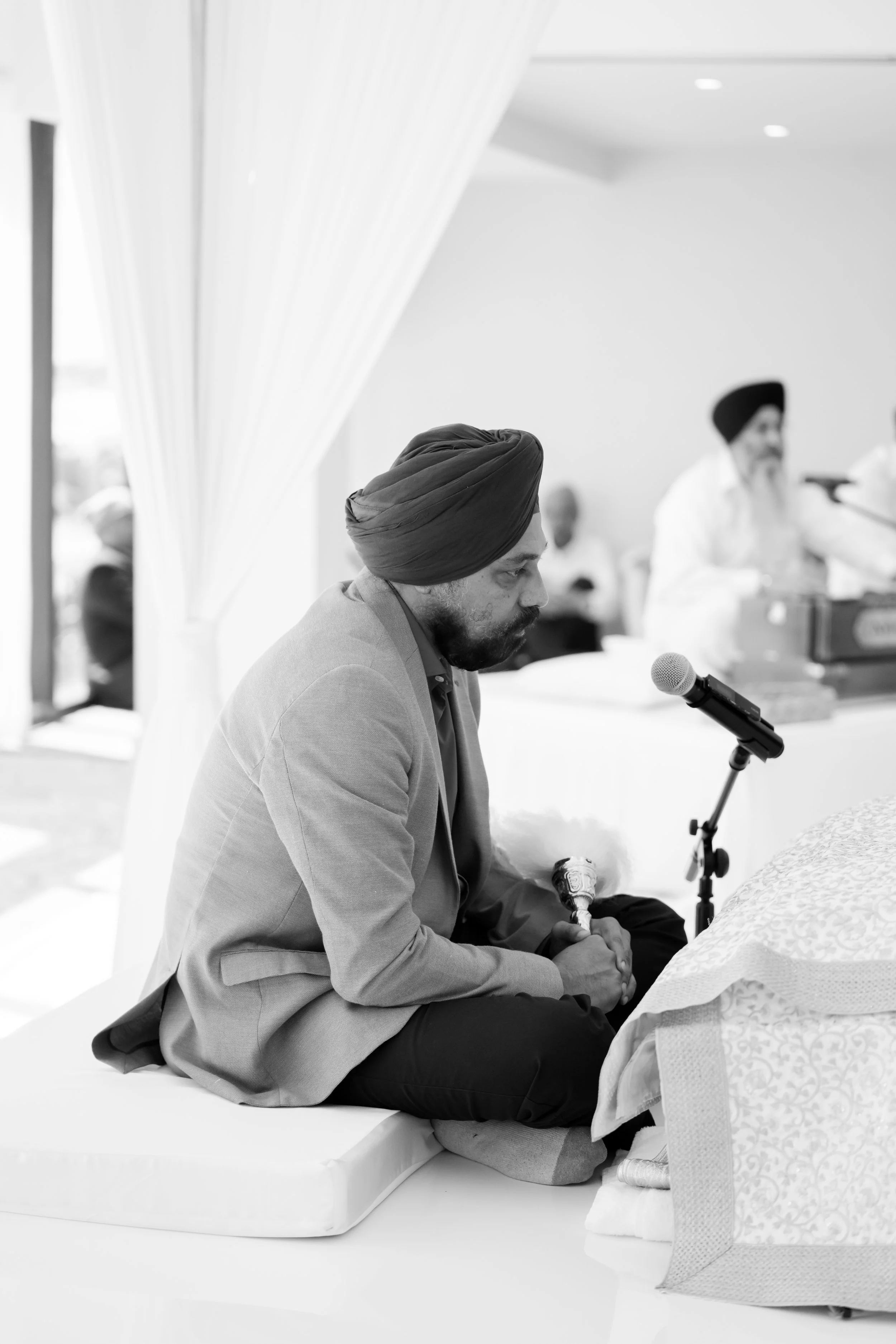 A man wearing a turban and suit sitting on a cushion, holding a ceremonial object, during a religious or cultural ceremony in a decorated room.