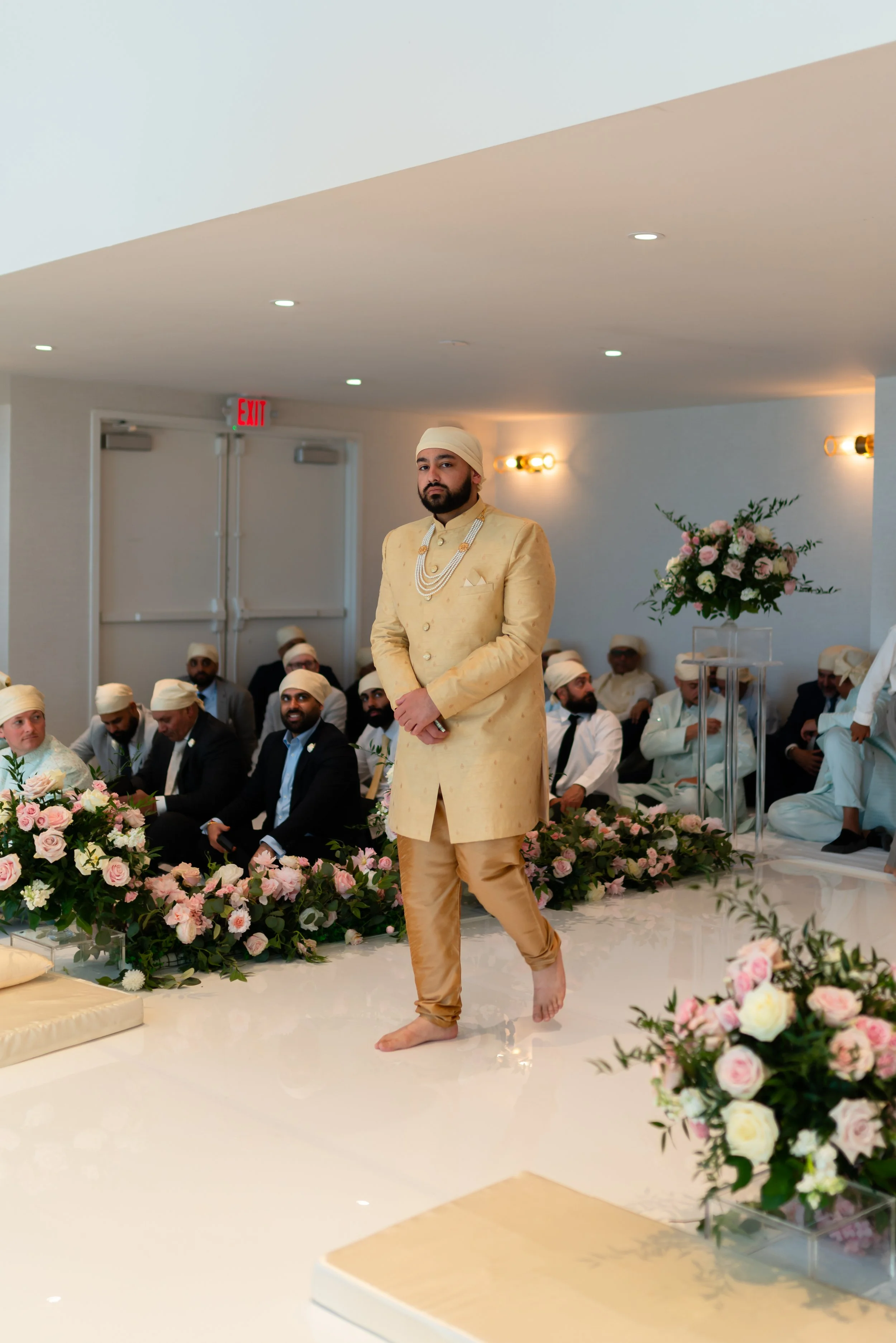 A groom in a cream-colored traditional Indian outfit with jewelry and a turban stands barefoot on a decorated stage at a wedding ceremony, with wedding guests seated in the background.