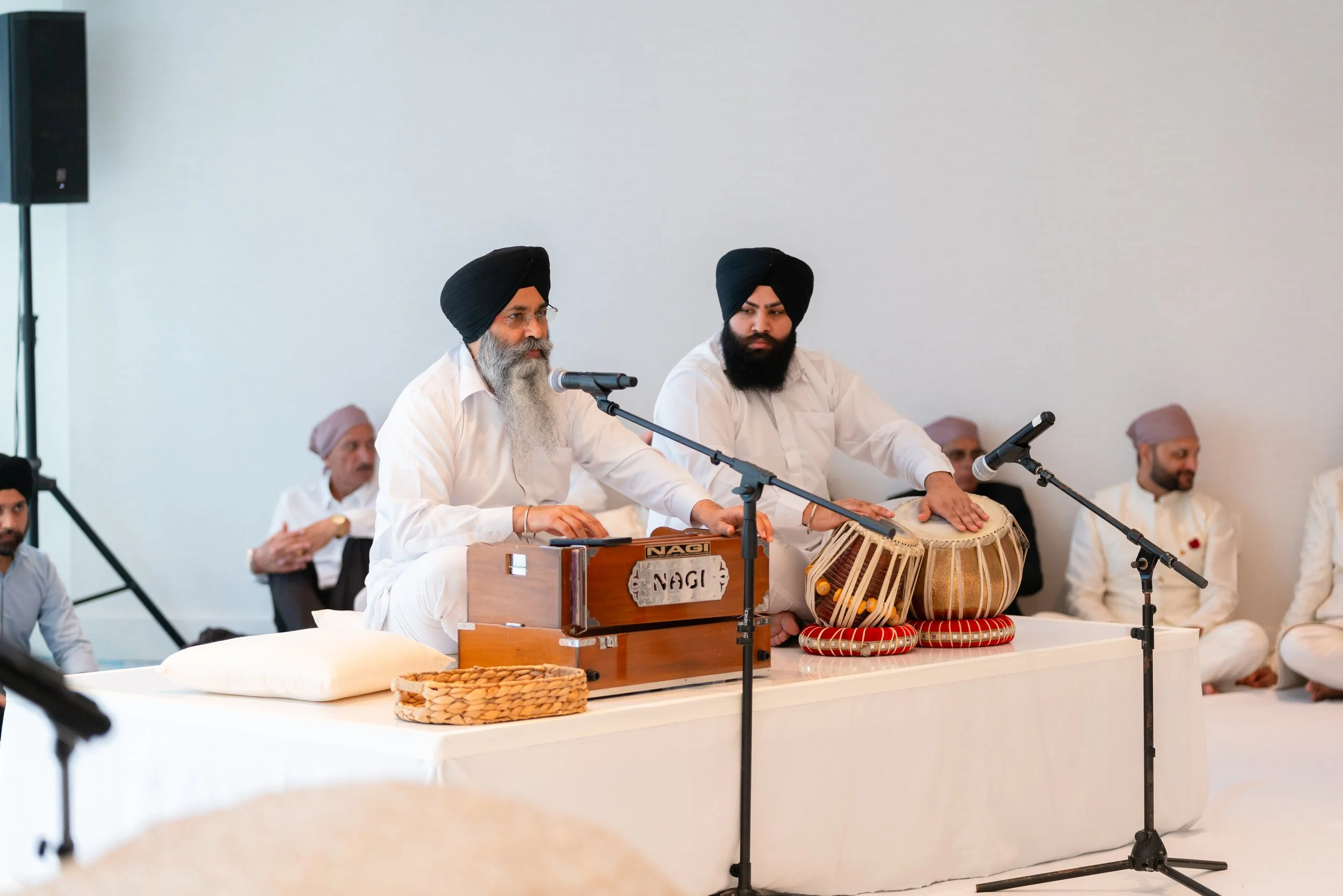 Two men wearing white kurtas and black turbans playing Punjabi musical instruments, with others in the background, seated on a stage during a cultural event.