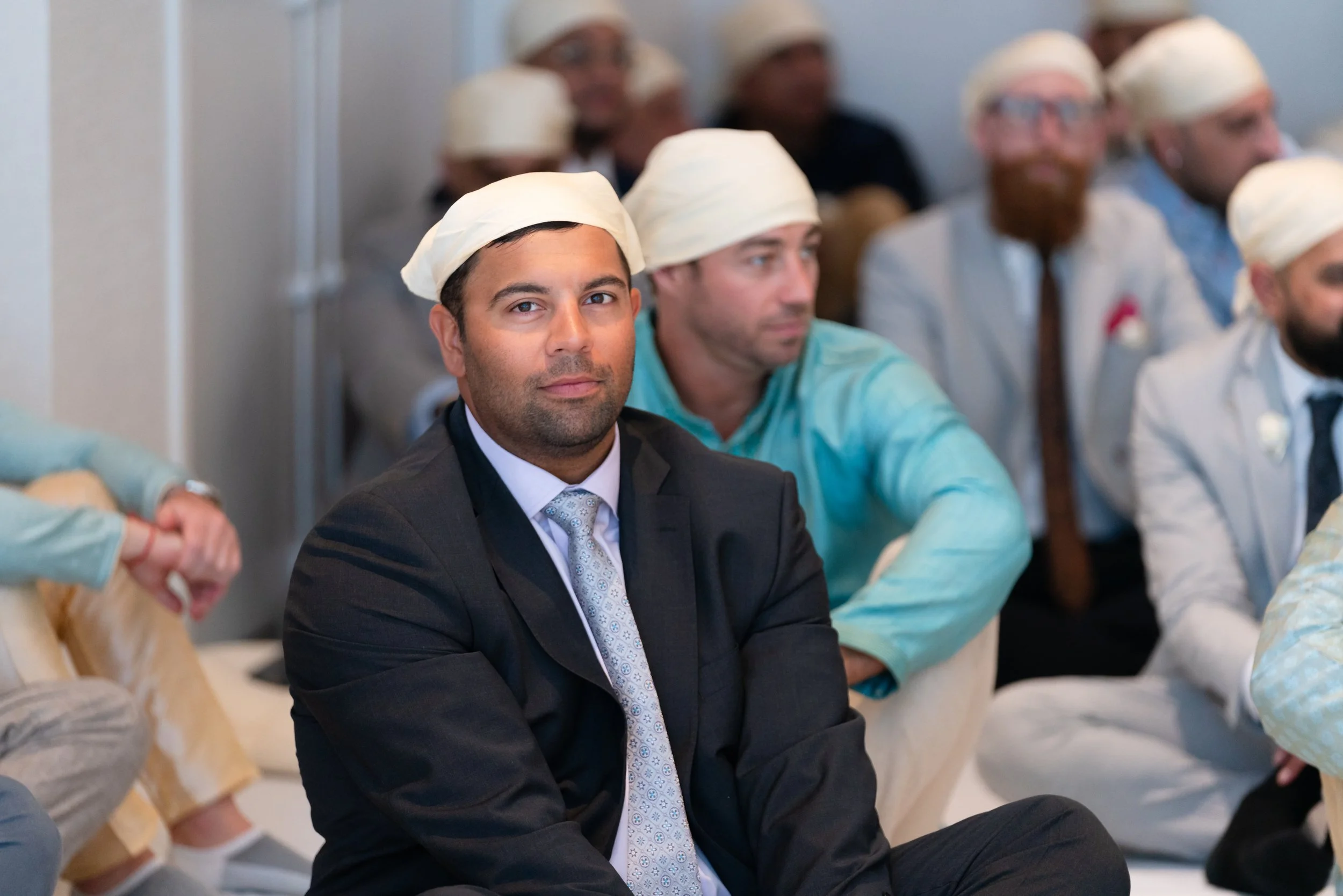 A man in a suit and tie with a white head covering sitting among other men in similar head coverings, attending a formal event or conference.