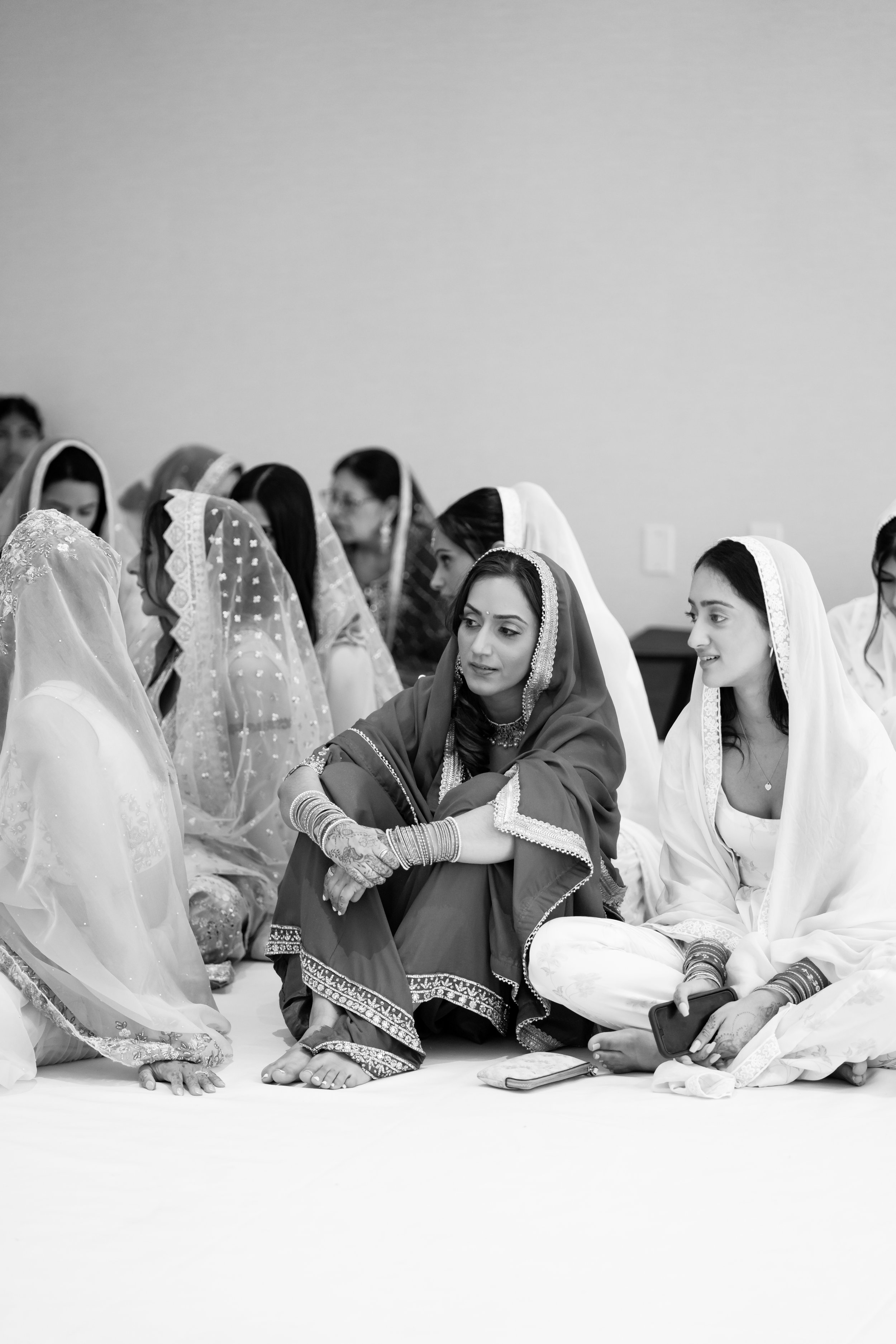 Group of women dressed in traditional South Asian attire, sitting on the floor in a cheery indoor setting.