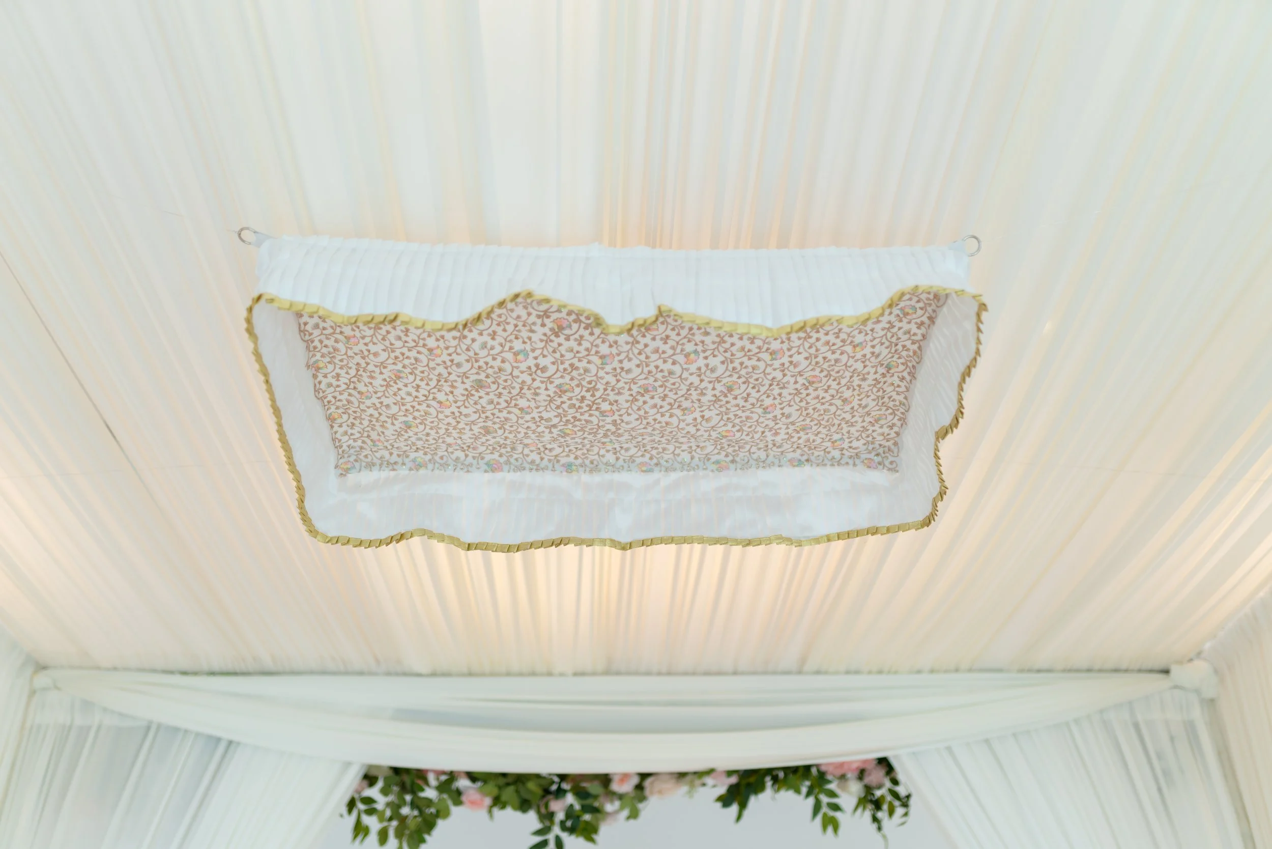 Decorative wall hanging with a floral pattern, framed by a white border with gold trim, mounted on a light-colored, pleated wall panel.