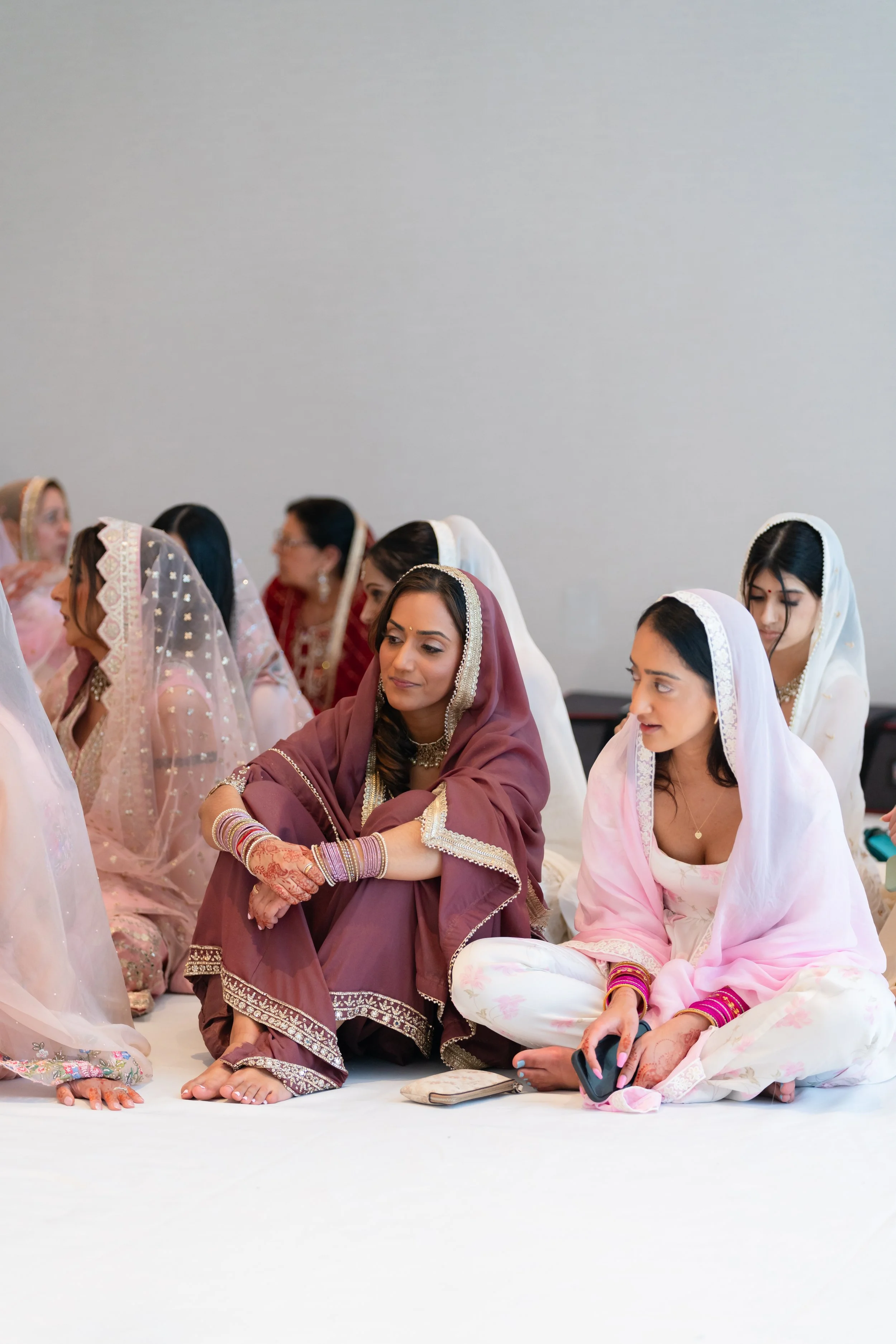 Group of women wearing traditional Indian sarees and dupattas, sitting on the floor during a cultural or religious event.
