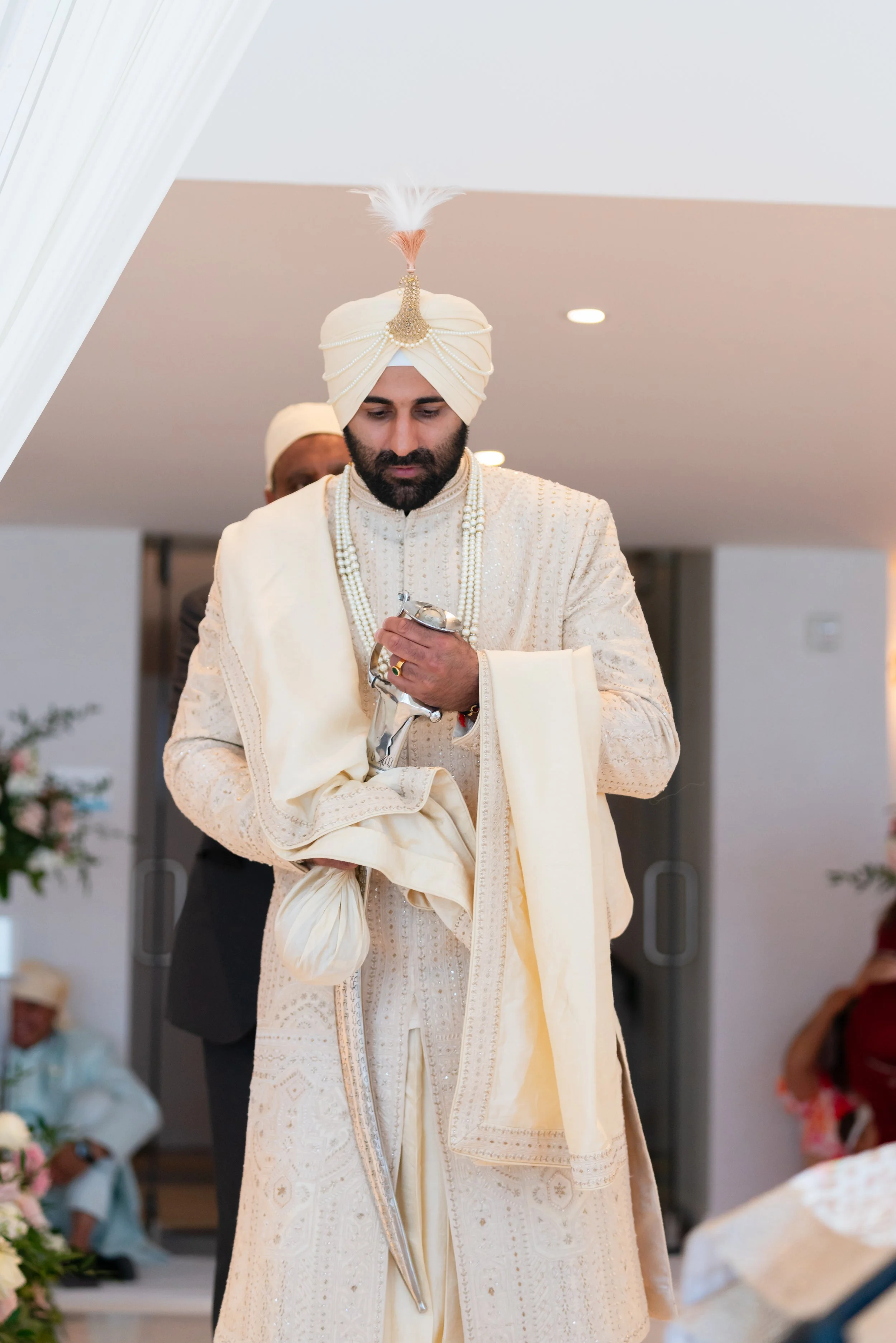 An Indian groom dressed in traditional cream-colored wedding attire, wearing a turban with a feather ornament, holding a folded cloth and a small silver object, during a wedding ceremony.