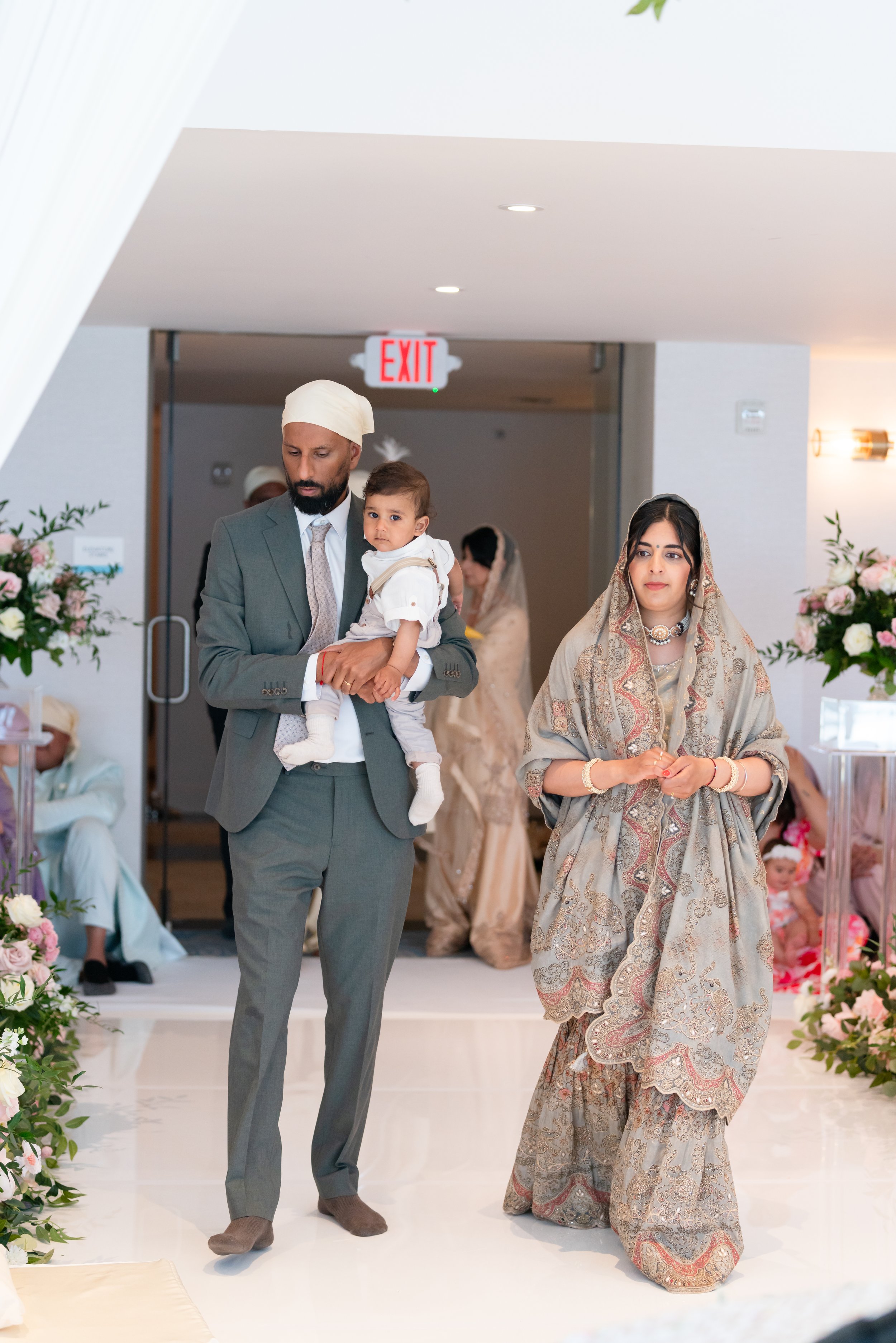 A man in a gray suit holding a young boy in traditional Indian attire, and a woman in traditional Indian clothing with jewelry, walking in a decorated indoor setting for a special occasion.