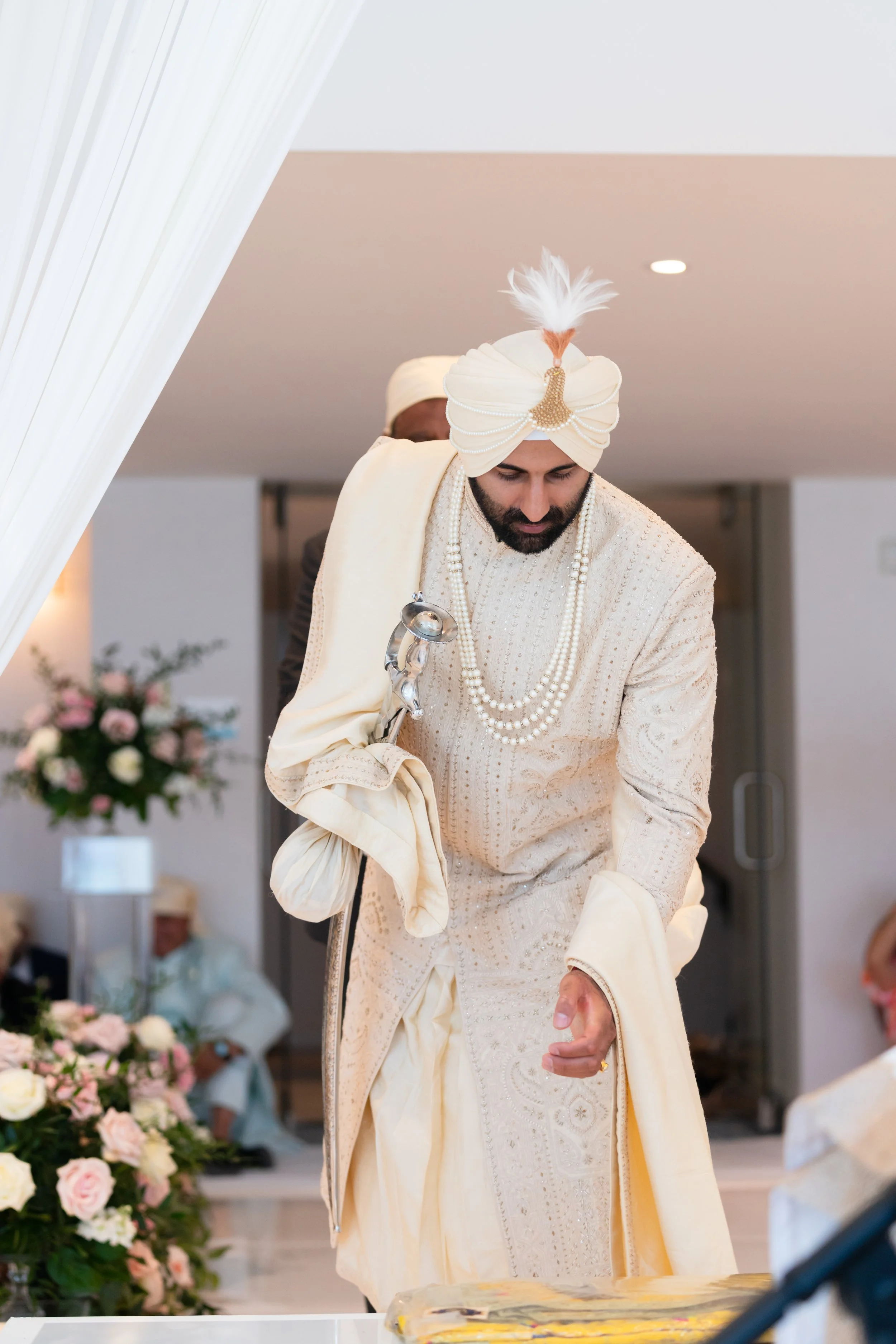 A groom dressed in traditional Indian wedding attire, including a cream-colored sherwani and a turban with a feather, at a wedding ceremony.
