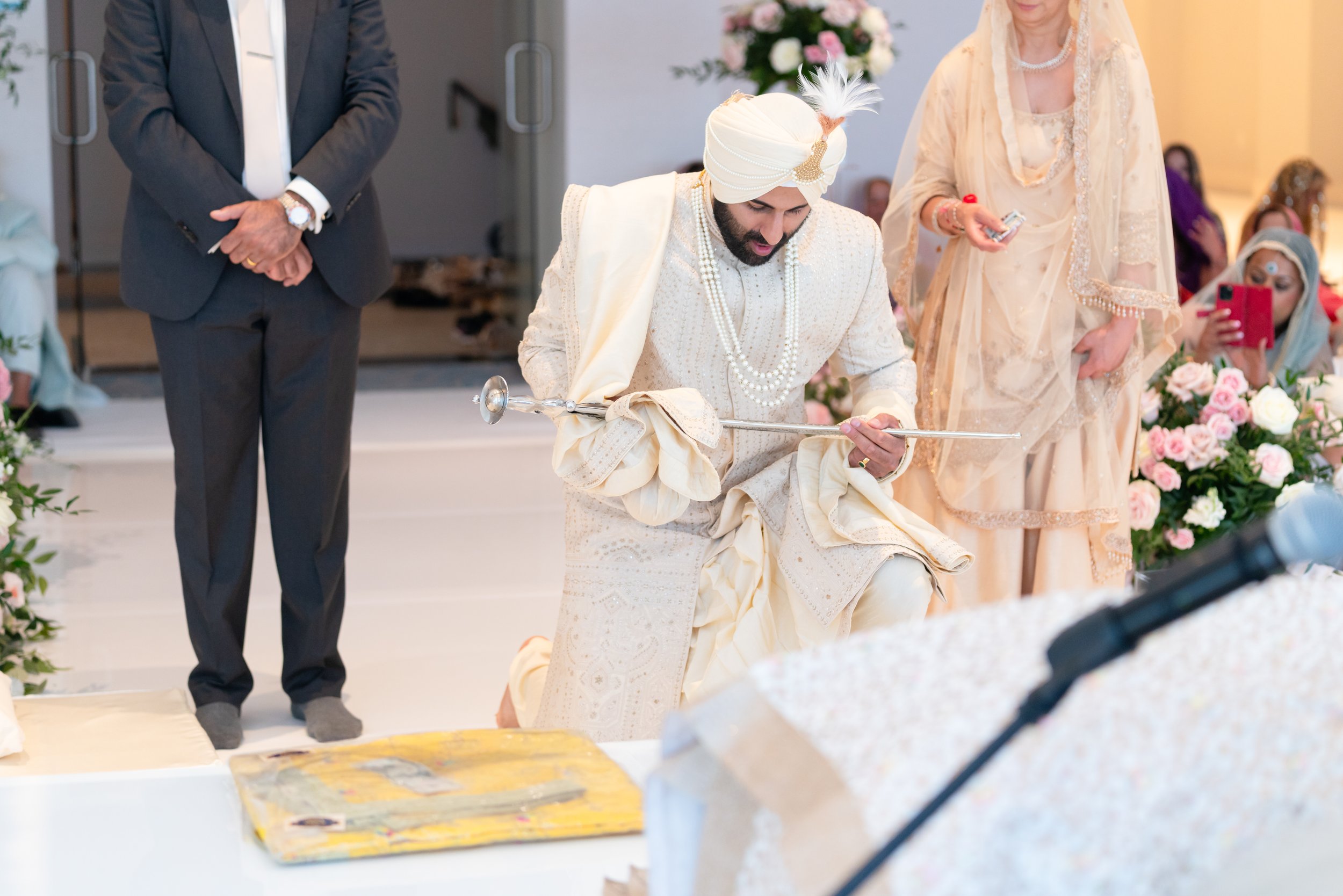 A man dressed in traditional Indian wedding attire kneels on one knee, holding a sword, during a wedding ceremony. He wears a cream-colored sherwani and turban with a feather. Several people stand around him, some taking photos. The setting is decora