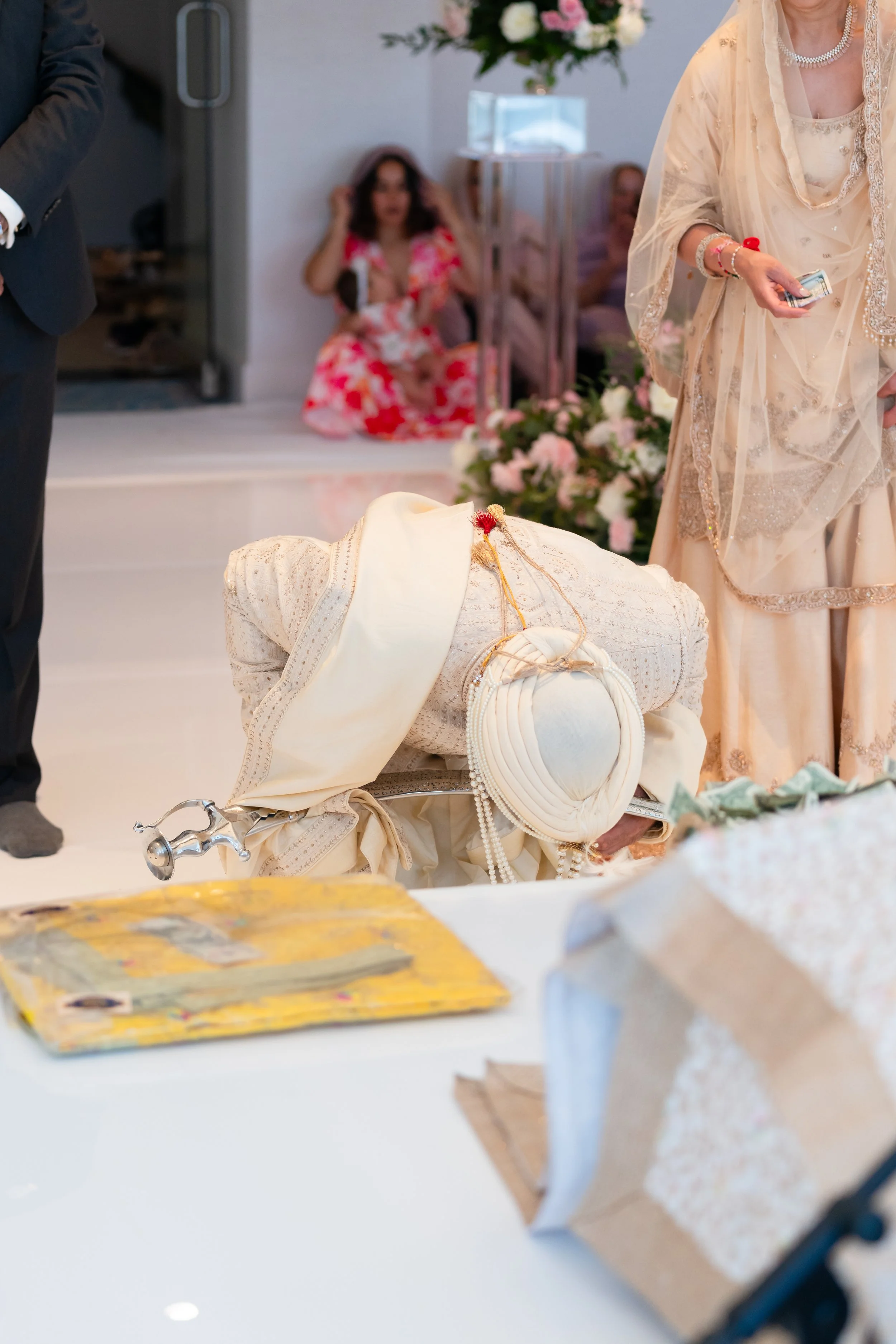 A person dressed in traditional attire is bowing or kneeling on the floor, with others standing around, during a ceremonial event. There are flowers and decorative items in the background.