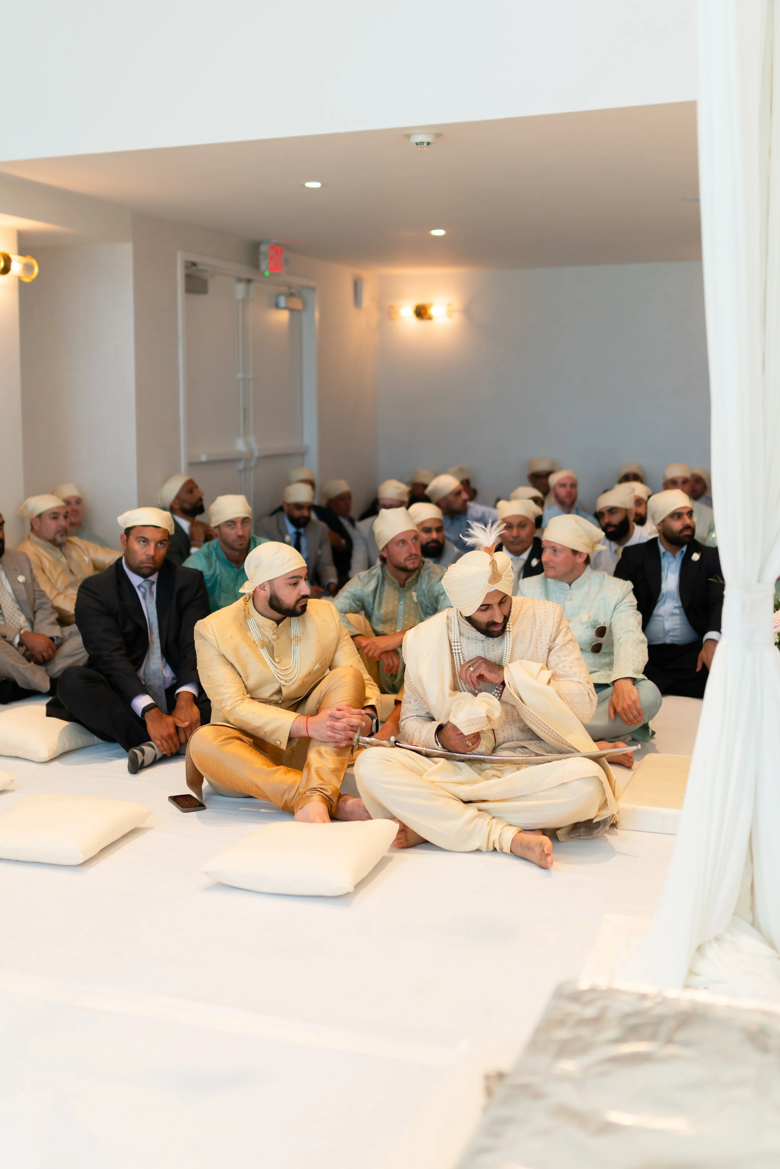 Indian wedding ceremony with groom and guests dressed in traditional attire, seated on the floor with white cushions.