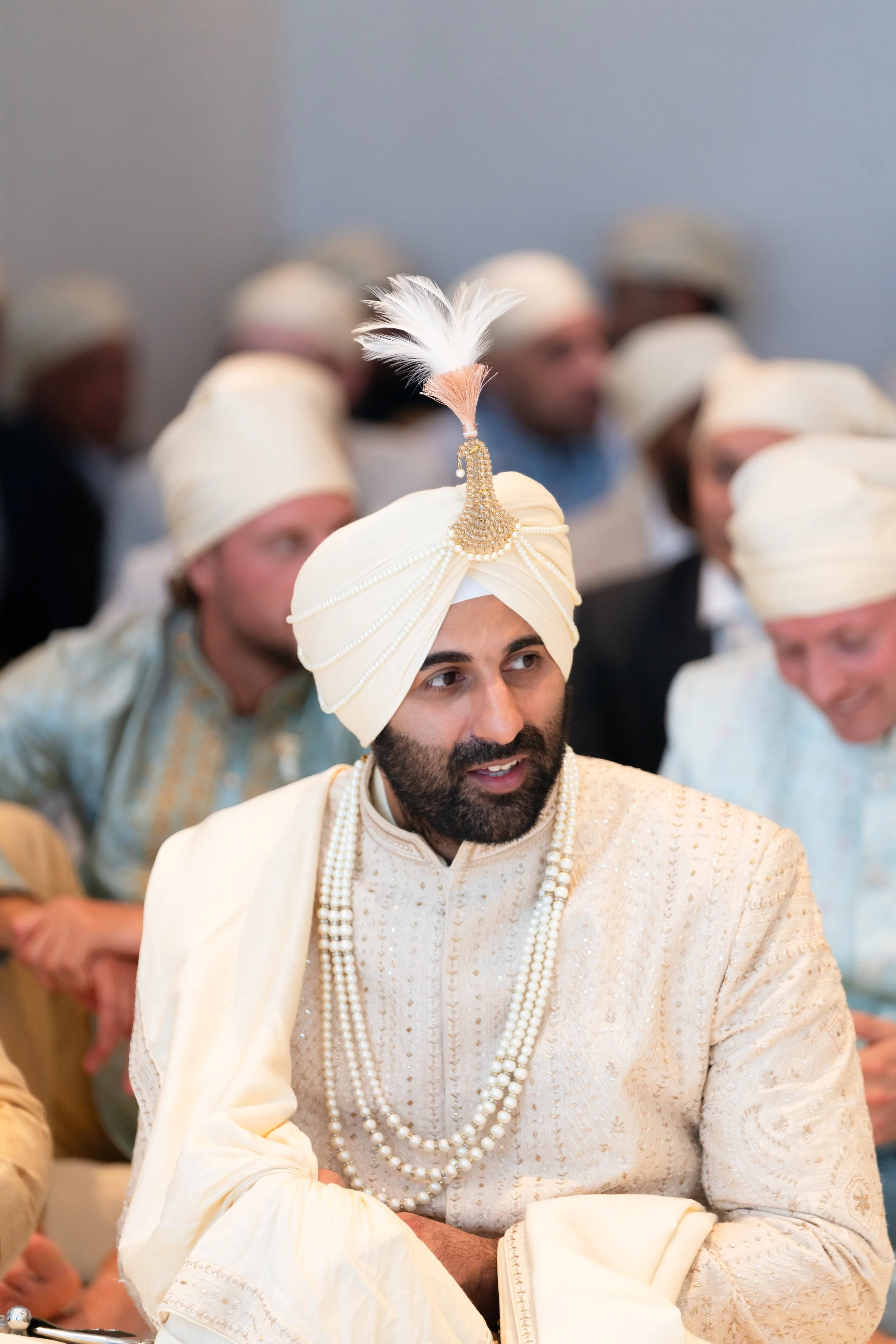 Man wearing a cream-colored turban with a decorative feather, pearl embellishments, and traditional Indian attire, surrounded by other men in similar attire.