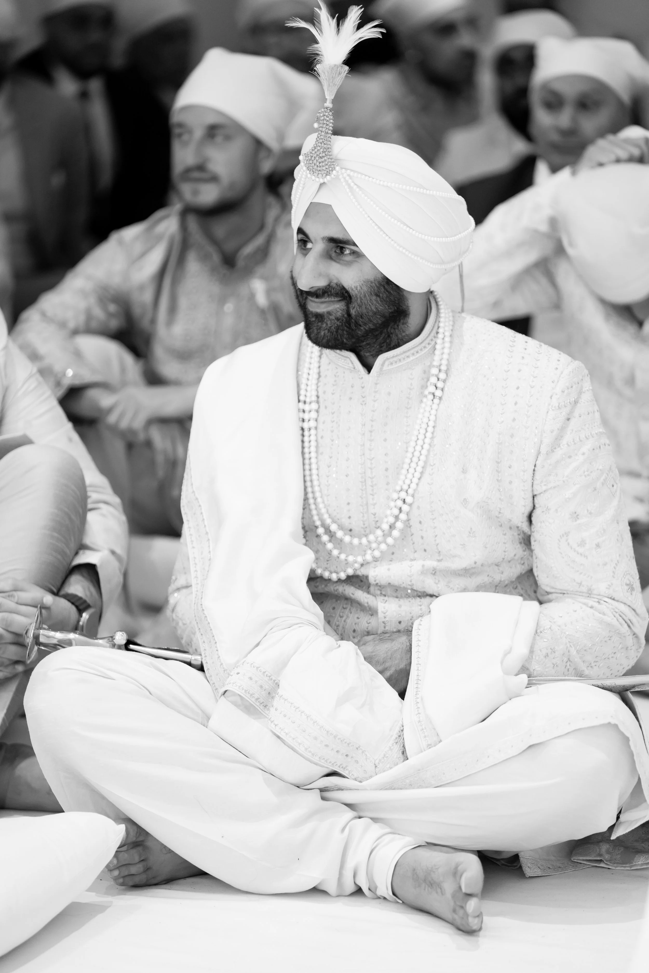 A man dressed in traditional Indian wedding attire, wearing a white sherwani, turban with a feather, and multiple pearl necklaces, seated cross-legged at a wedding ceremony.