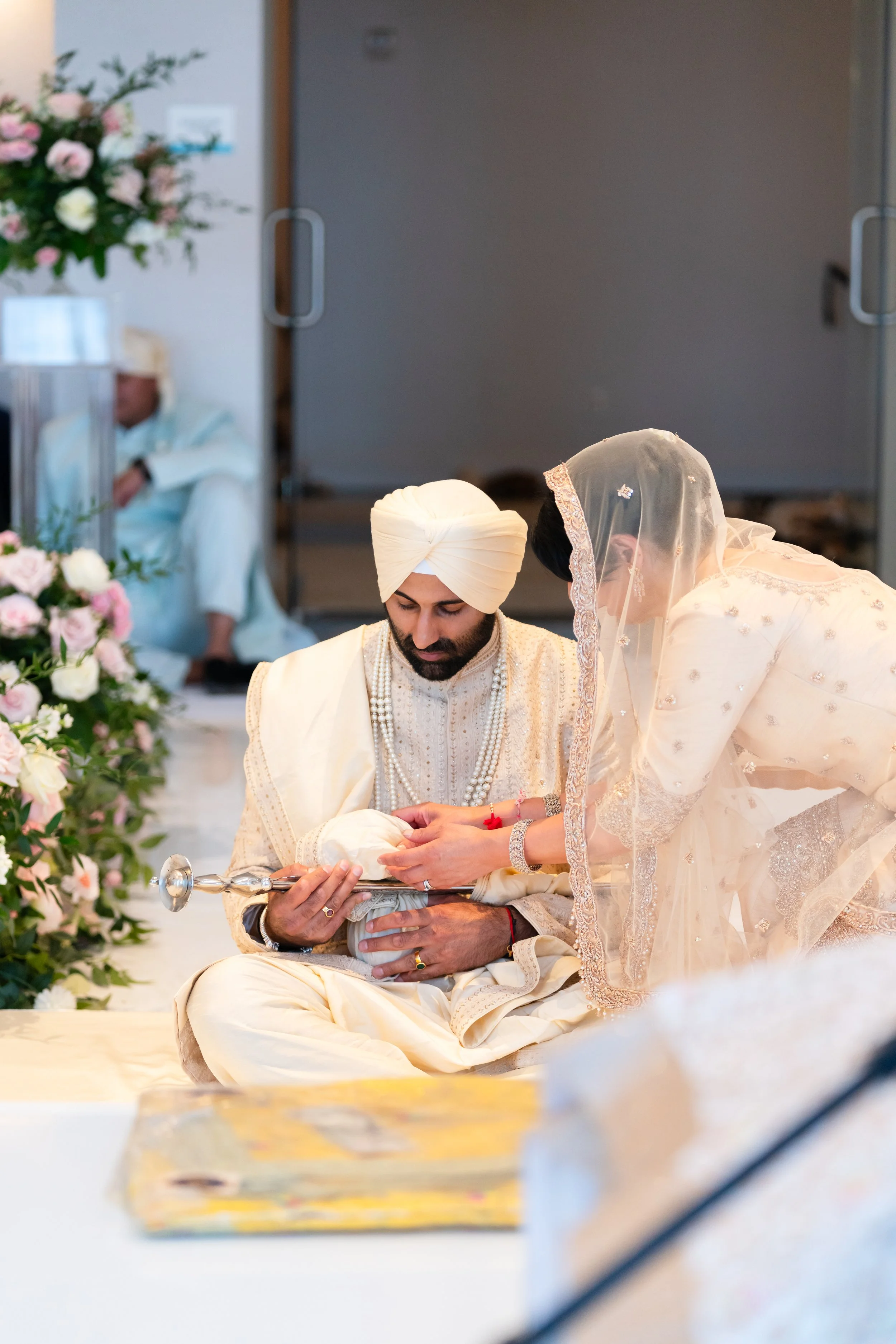 A traditional Indian wedding ceremony with a bride and groom dressed in cream-colored wedding attire, sitting on the floor, with the bride holding a ceremonial sword, surrounded by floral decorations.
