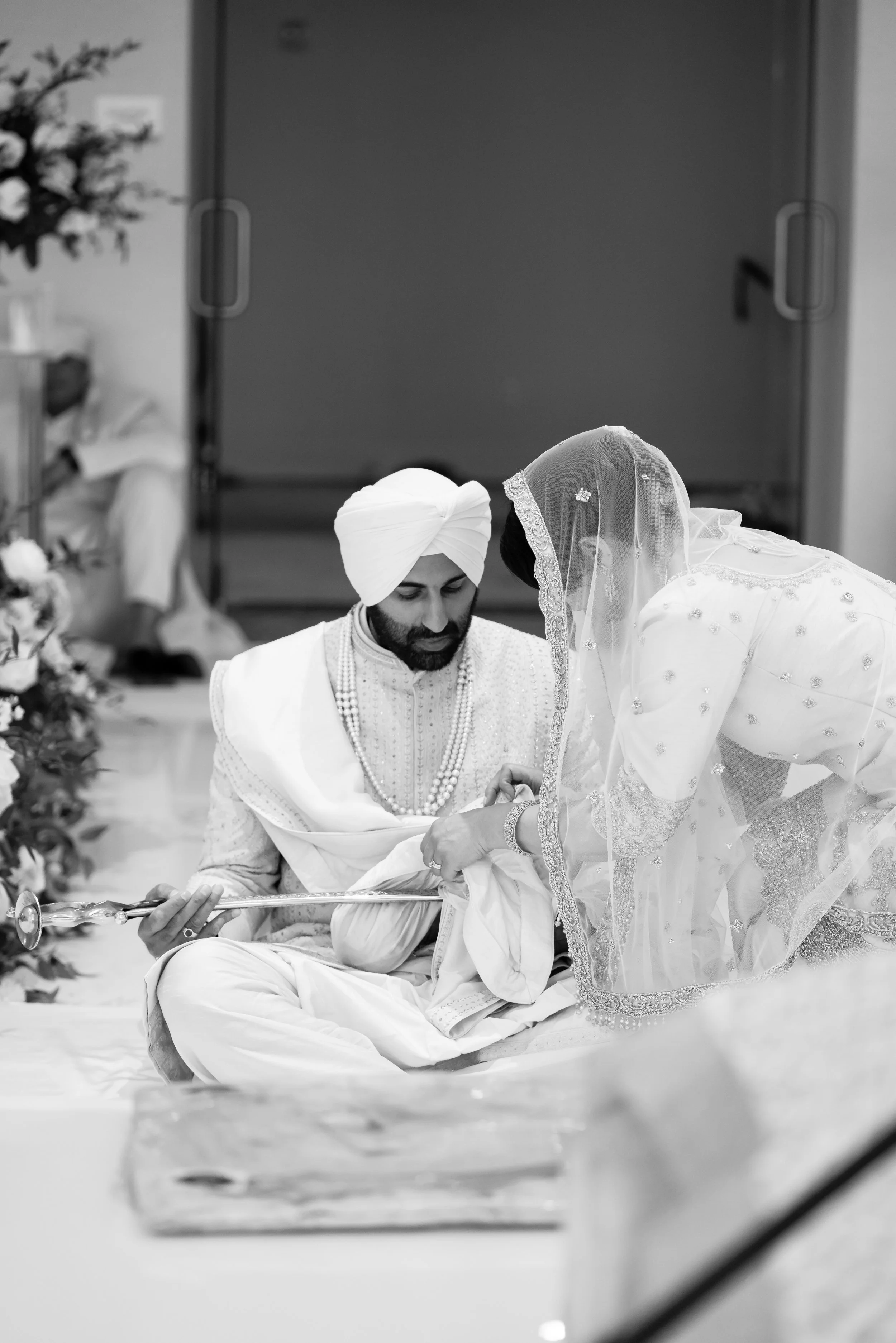 A traditional Indian wedding ceremony with a groom in a turban and a bride in a saree, both dressed in ornate attire, participating in a ritual involving a sword.