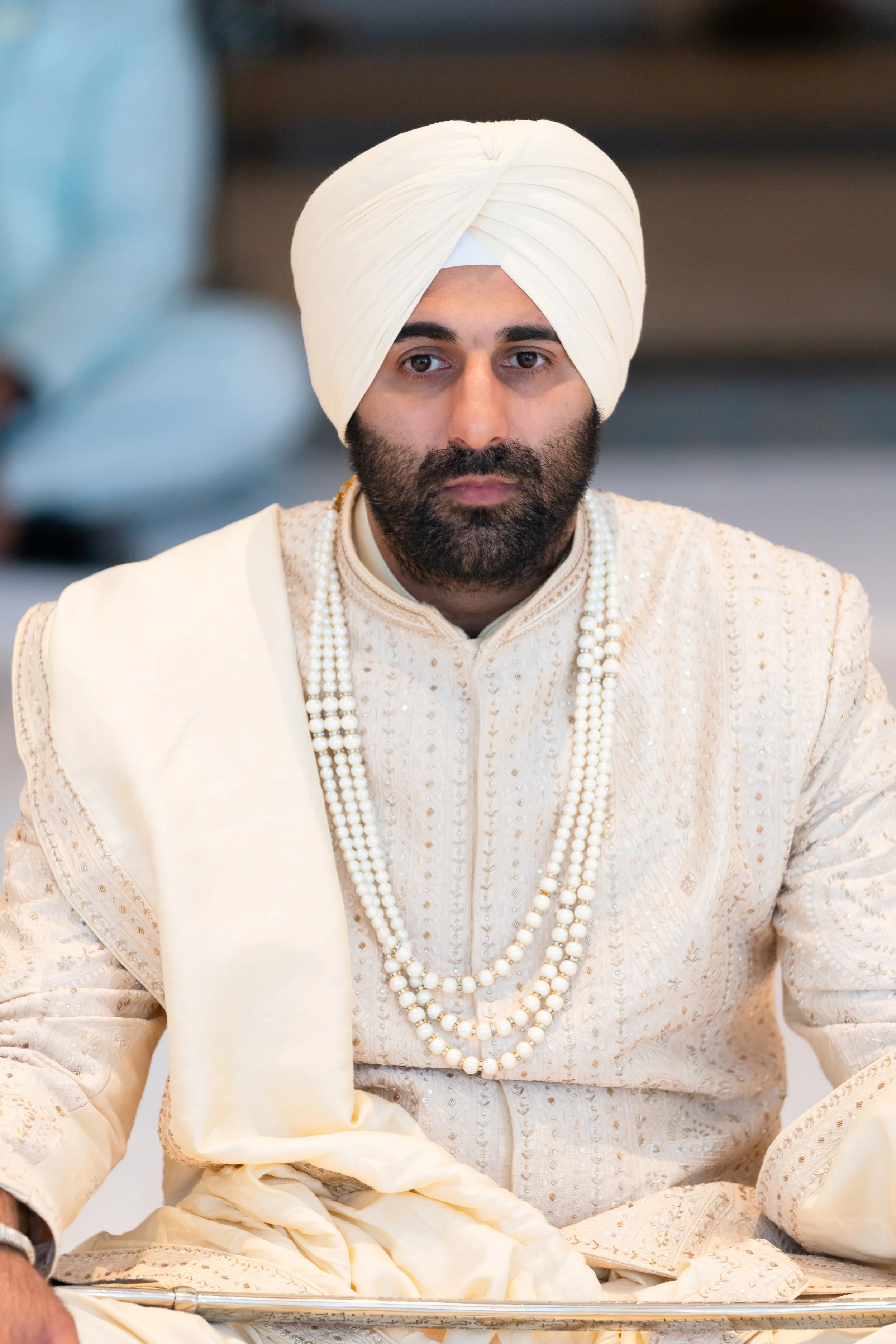 A man in traditional Indian wedding attire, wearing a cream-colored sherwani with intricate embroidery, multiple layers of pearl necklaces, and a cream turban, sitting with a serious expression.