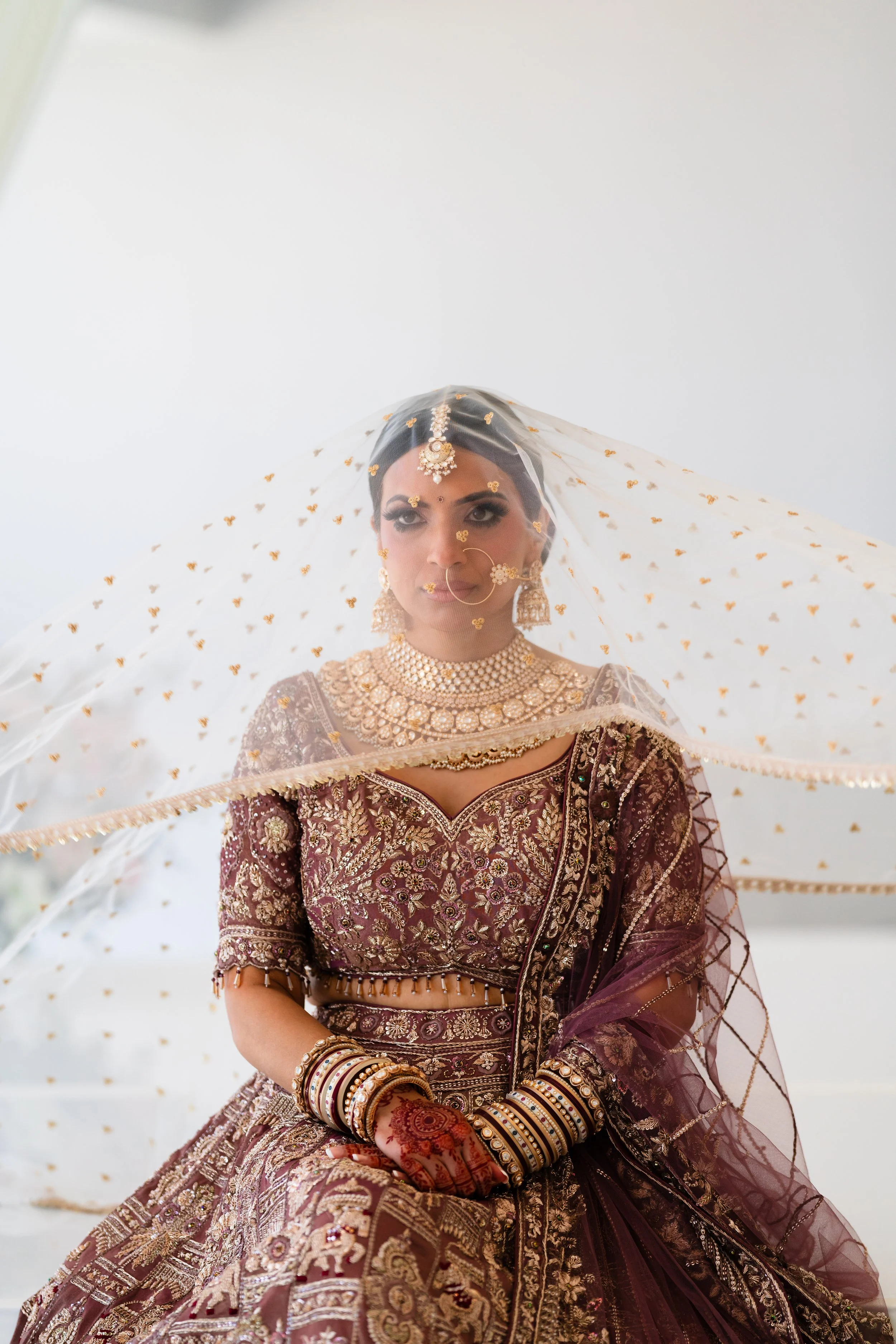 A bride dressed in an ornate maroon and gold traditional Indian wedding outfit, adorned with jewelry, henna, and a veil, sitting with hands folded.