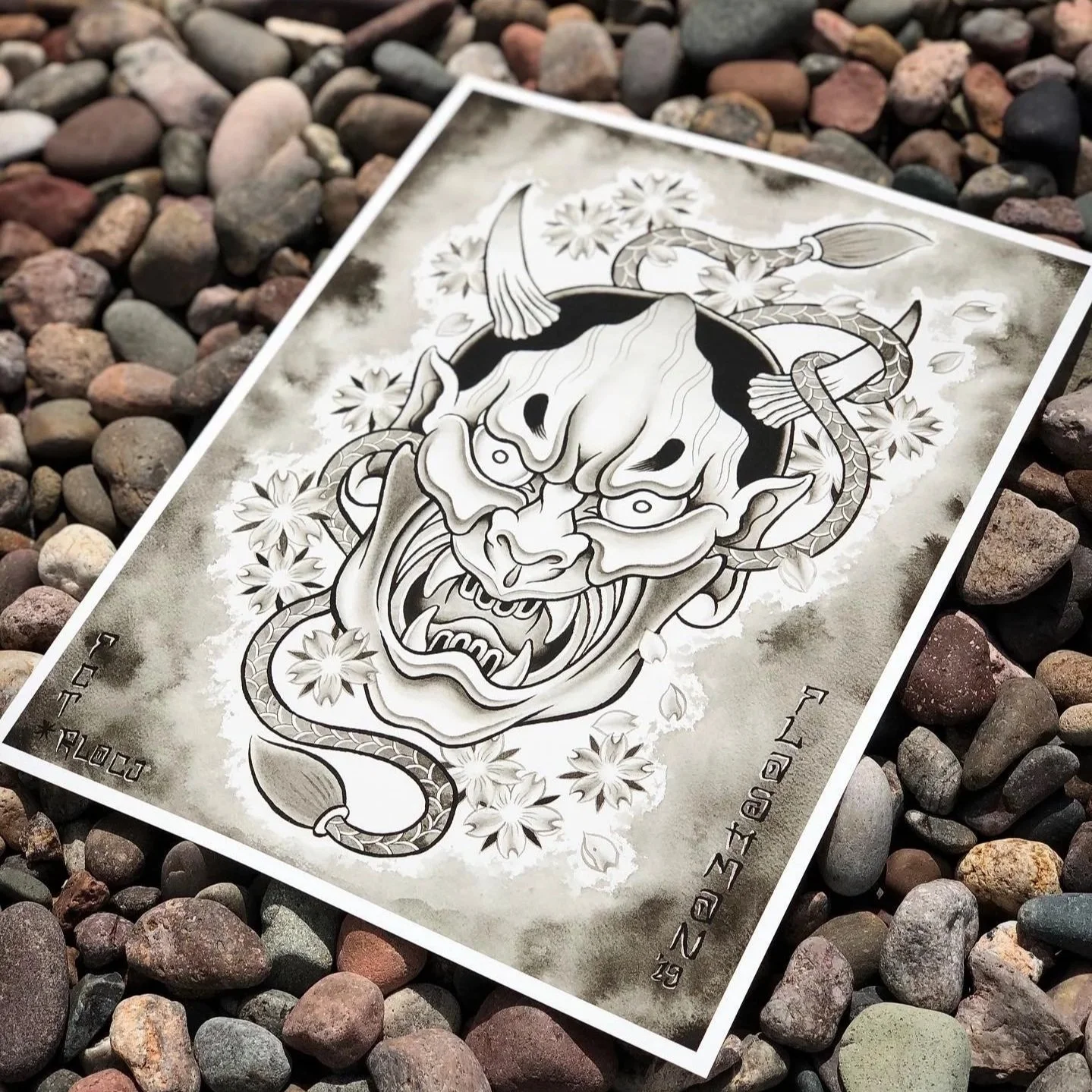 A black and white tattoo-style illustration of a fierce demon or oni with a snake wrapped around its head, surrounded by cherry blossom flowers, on a piece of paper laying on multicolored rocks.