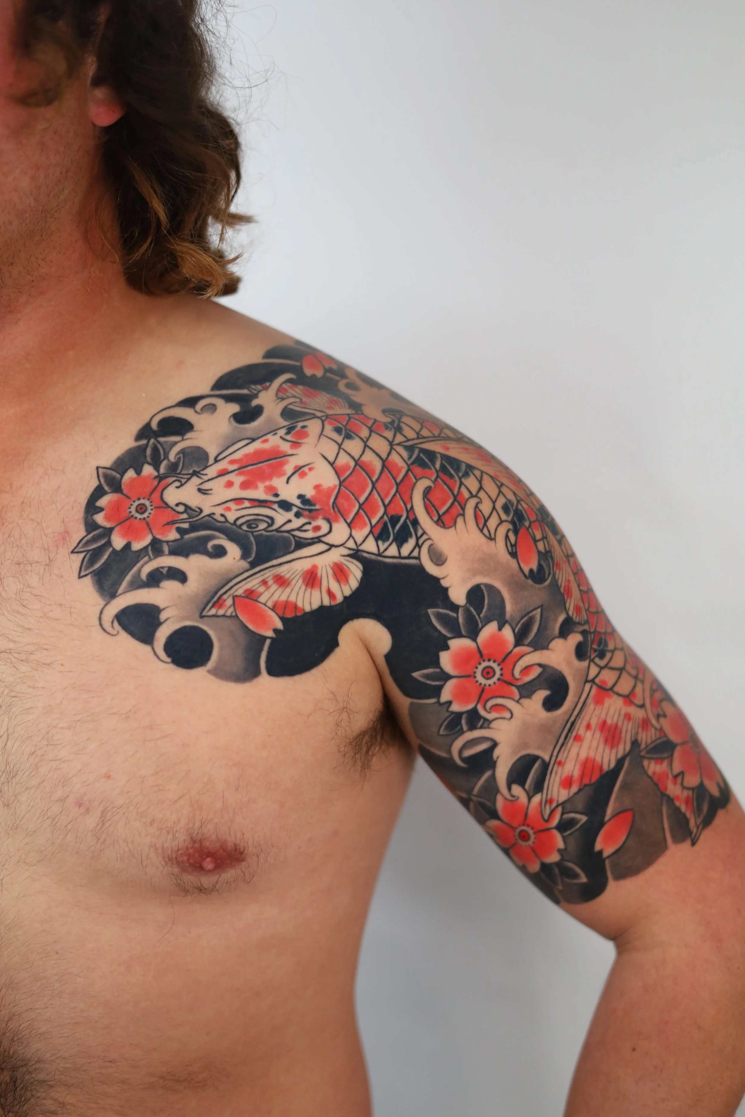 A detailed Japanese-style tattoo featuring a koi fish surrounded by cherry blossoms and black waves on a man's shoulder and upper arm.