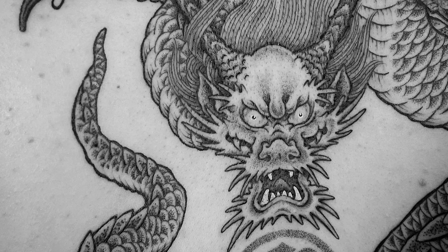 A detailed black and gray tattoo of a fierce dragon with a lion-like face, sharp eyes, and an open mouth showing fangs. The dragon has textured scales and a long, curved tail.