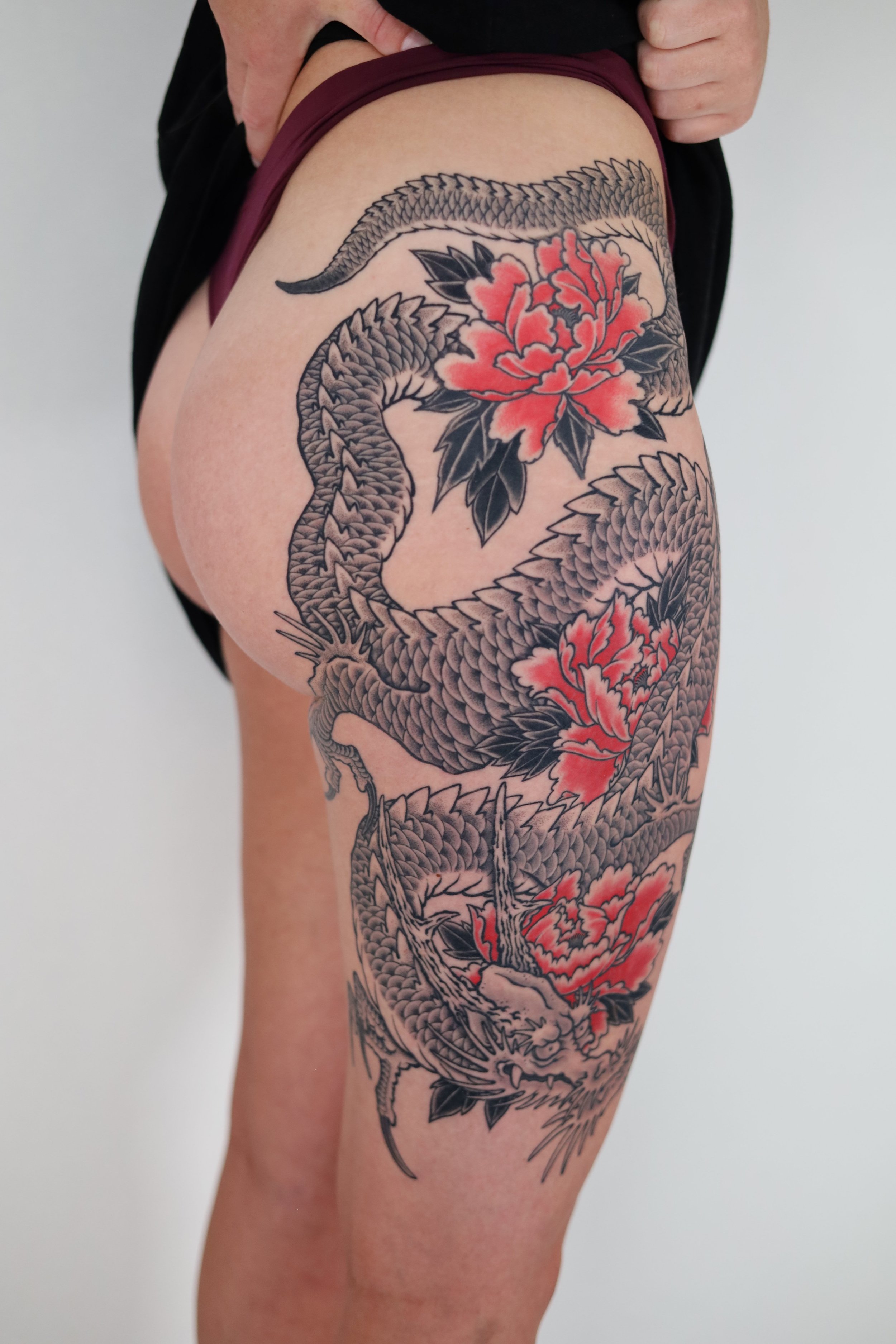A detailed black and gray tattoo of a dragon intertwined with red and black peony flowers on a person's thigh and hip.