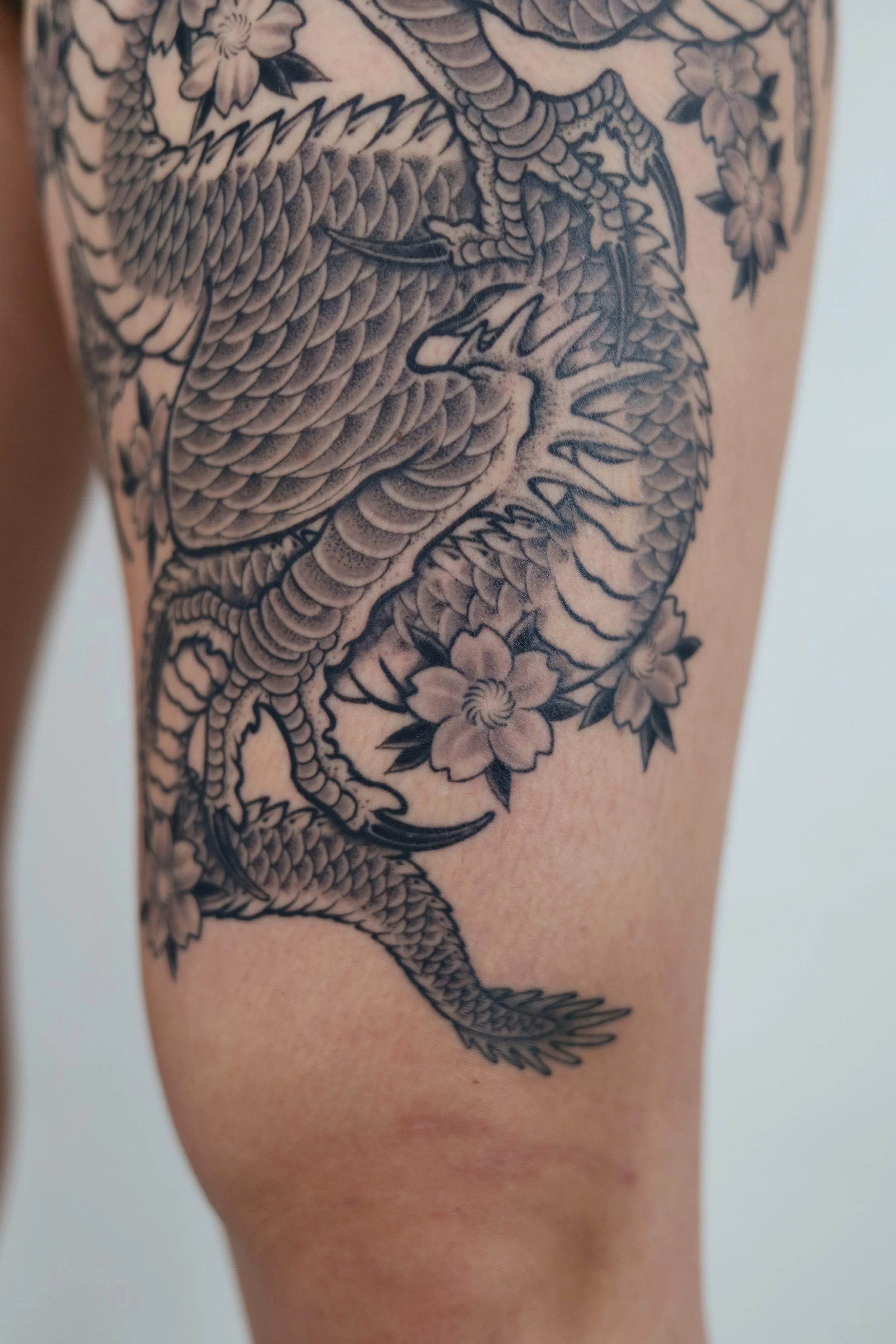 Close-up of a person's thigh featuring a detailed black and gray tattoo of a dragon intertwined with flowers.