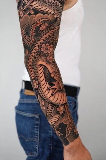 Close-up of a person's arm with an intricate black and gray traditional Japanese-style tattoo featuring a dragon, flowers, and waves.