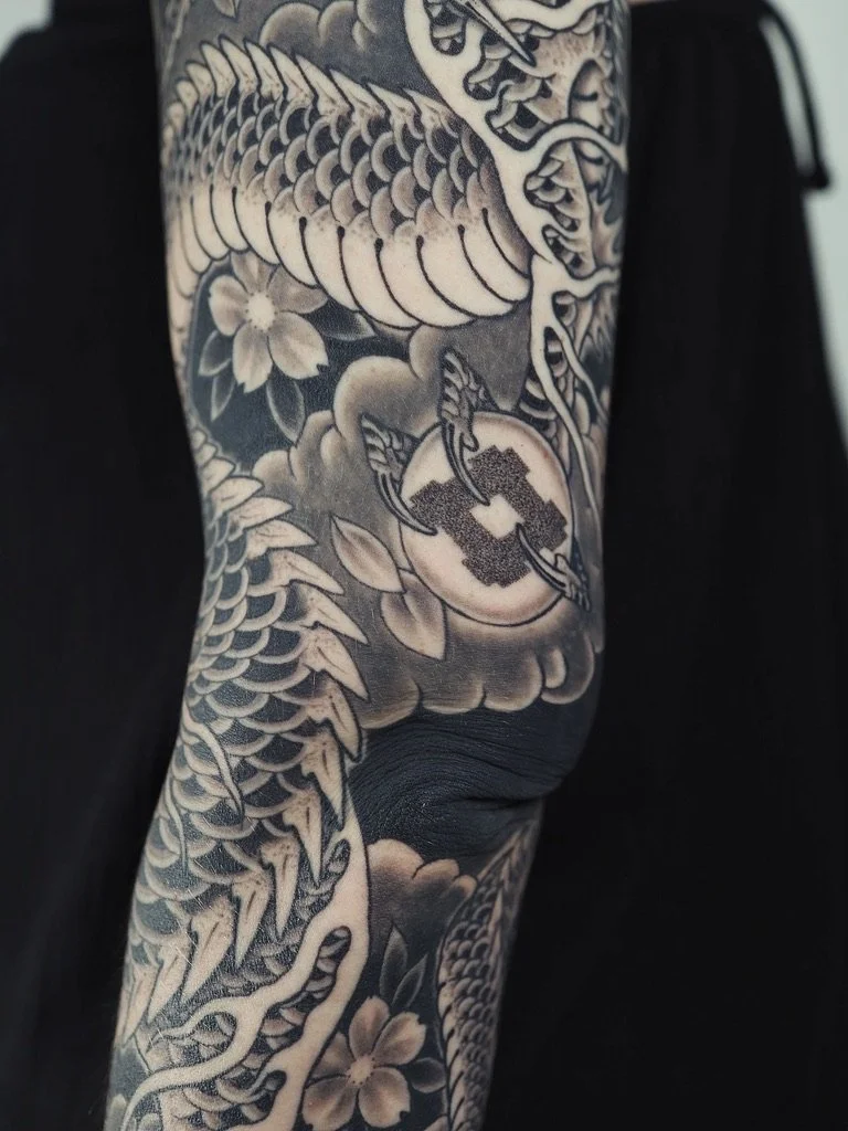 Black and gray Japanese-style tattoo sleeve featuring a dragon, a cherry blossom, and a circular emblem with a chain and a hook.