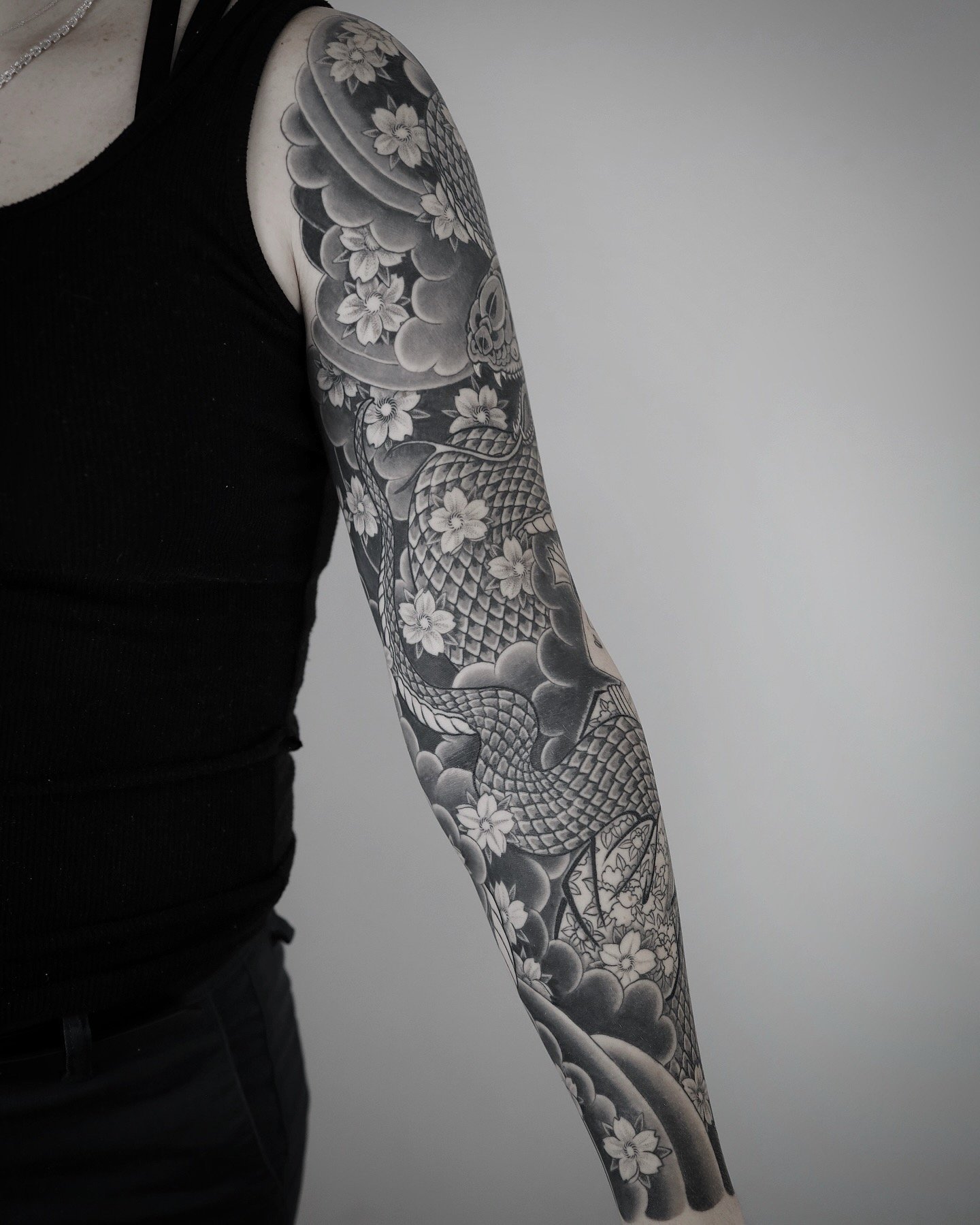 A person's arm with an intricate black and gray tattoo featuring a snake, flowers, clouds, and a skull.