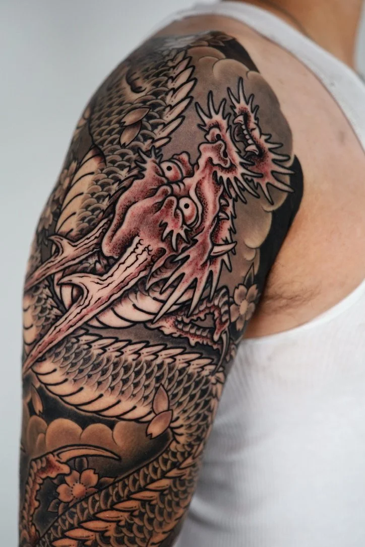 Close-up of a colorful dragon tattoo on a person's upper arm and shoulder, featuring detailed scales, a fierce face, and floral elements.