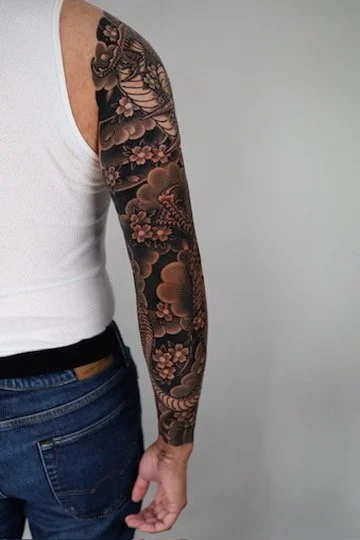 Close-up of a person wearing a white tank top and blue jeans, showing an intricate full-arm tattoo with floral and traditional Japanese imagery.