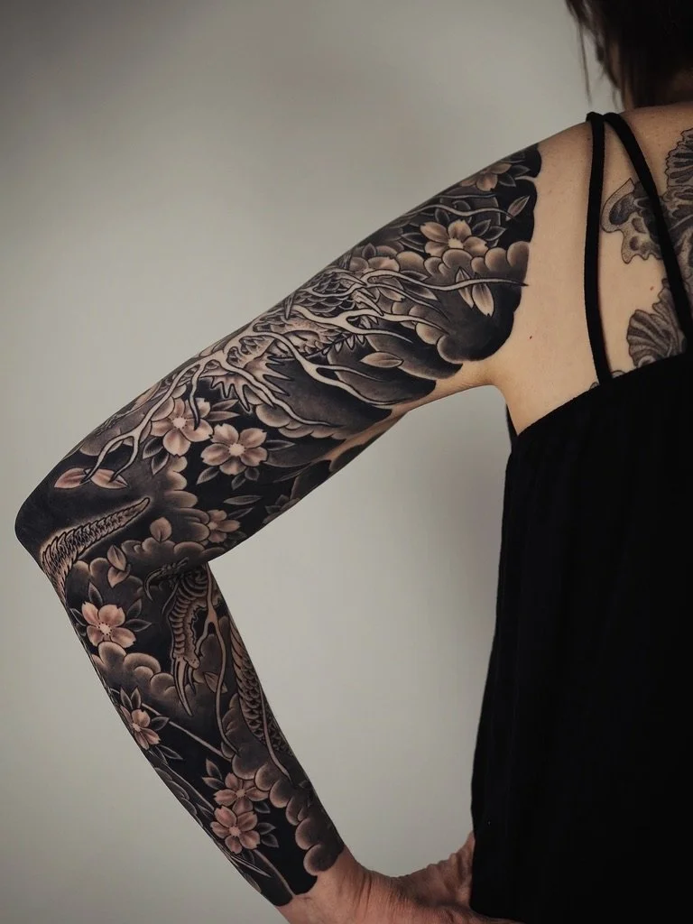 A person's arm and shoulder with a detailed black and gray tattoo sleeve featuring flowers and a snake.