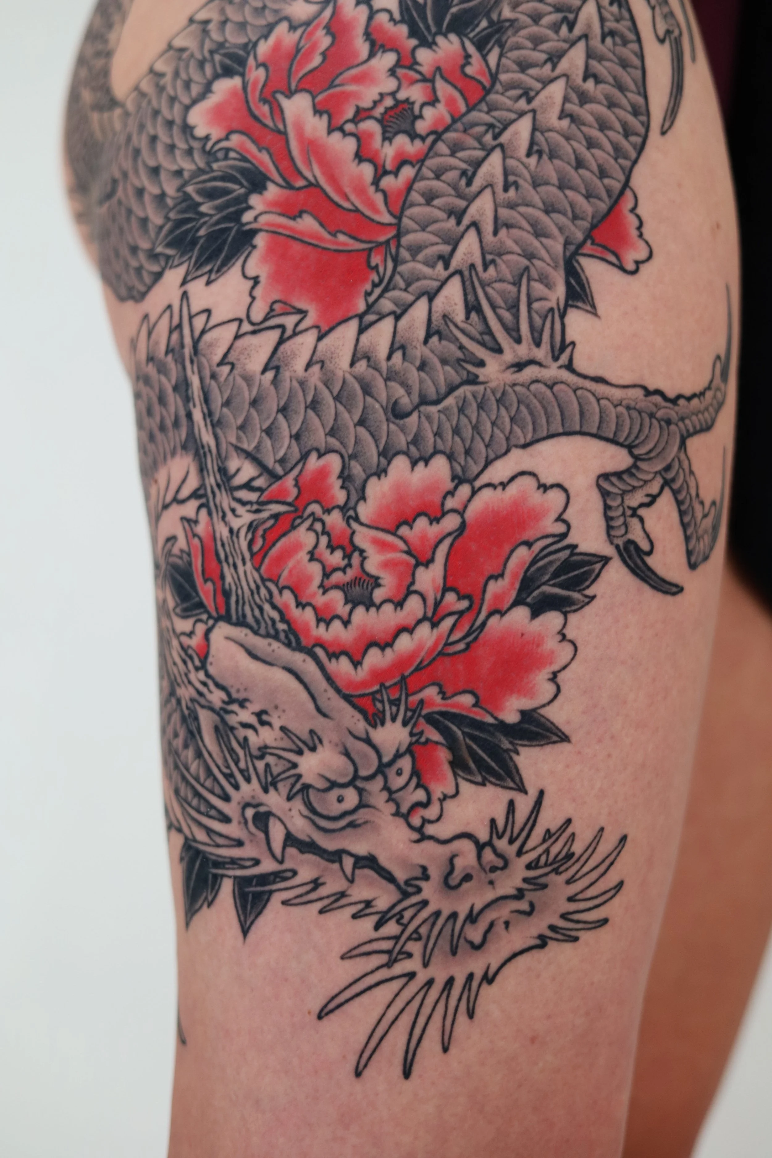Black and gray traditional style tattoo of a fierce dragon with sharp claws and scales, intertwined with large red and black peony flowers, covering the upper arm.