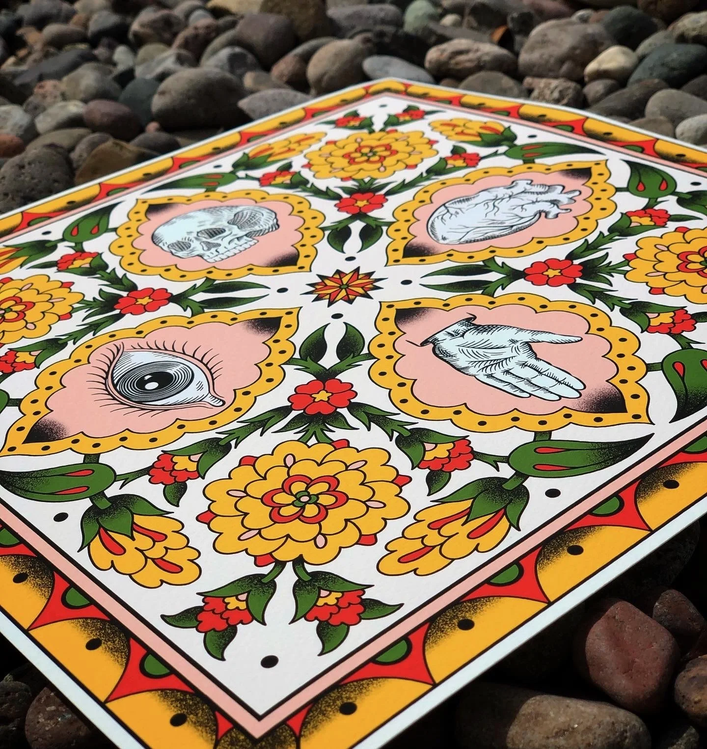 A decorative card with traditional Mexican artwork featuring colorful floral patterns and symbolic images including a skull, eye, heart, and hand, placed on a rocky surface.