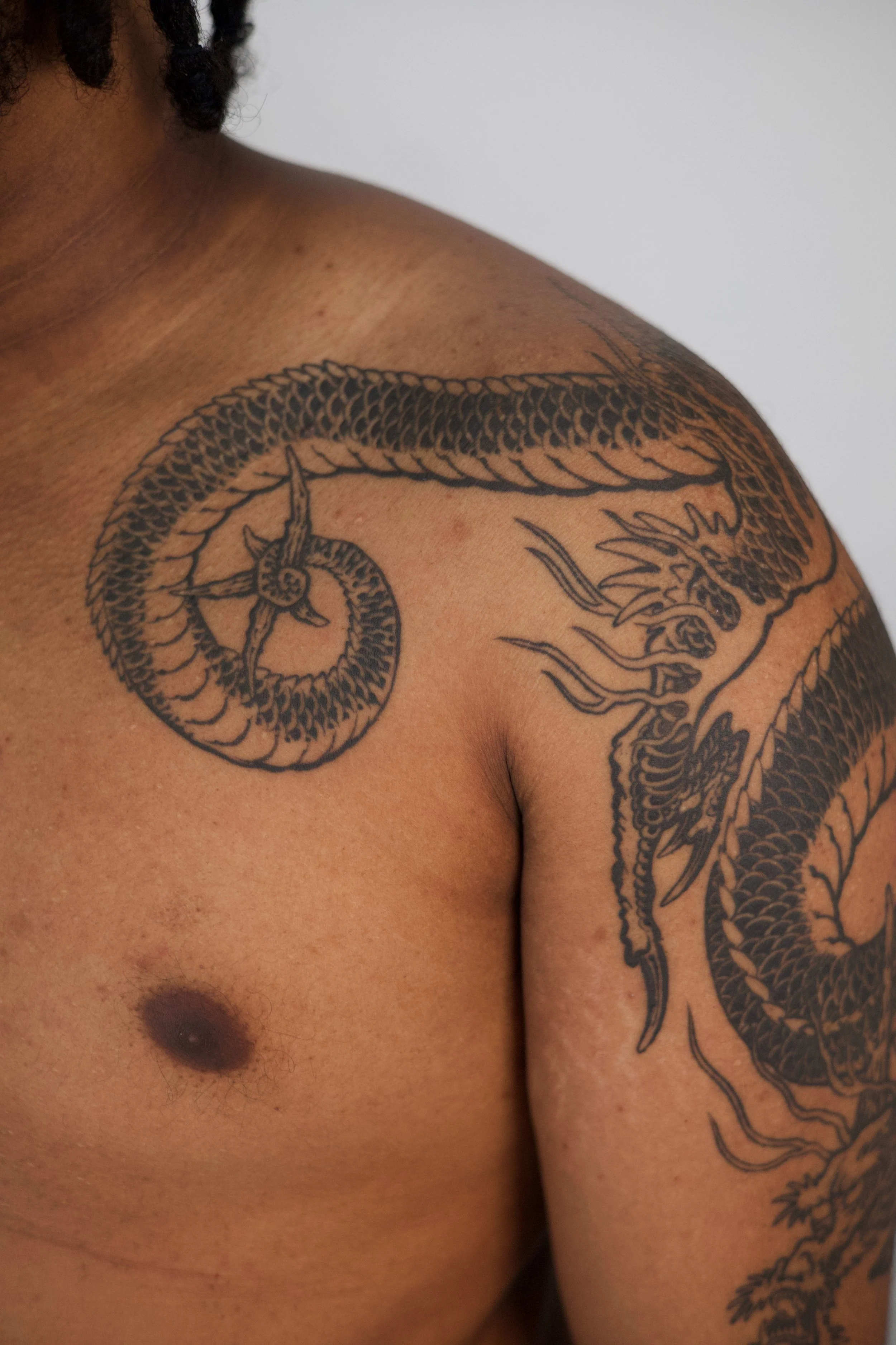 Close-up of a person's shoulder and chest with a detailed black tattoo of a dragon wrapping around the shoulder and arm.