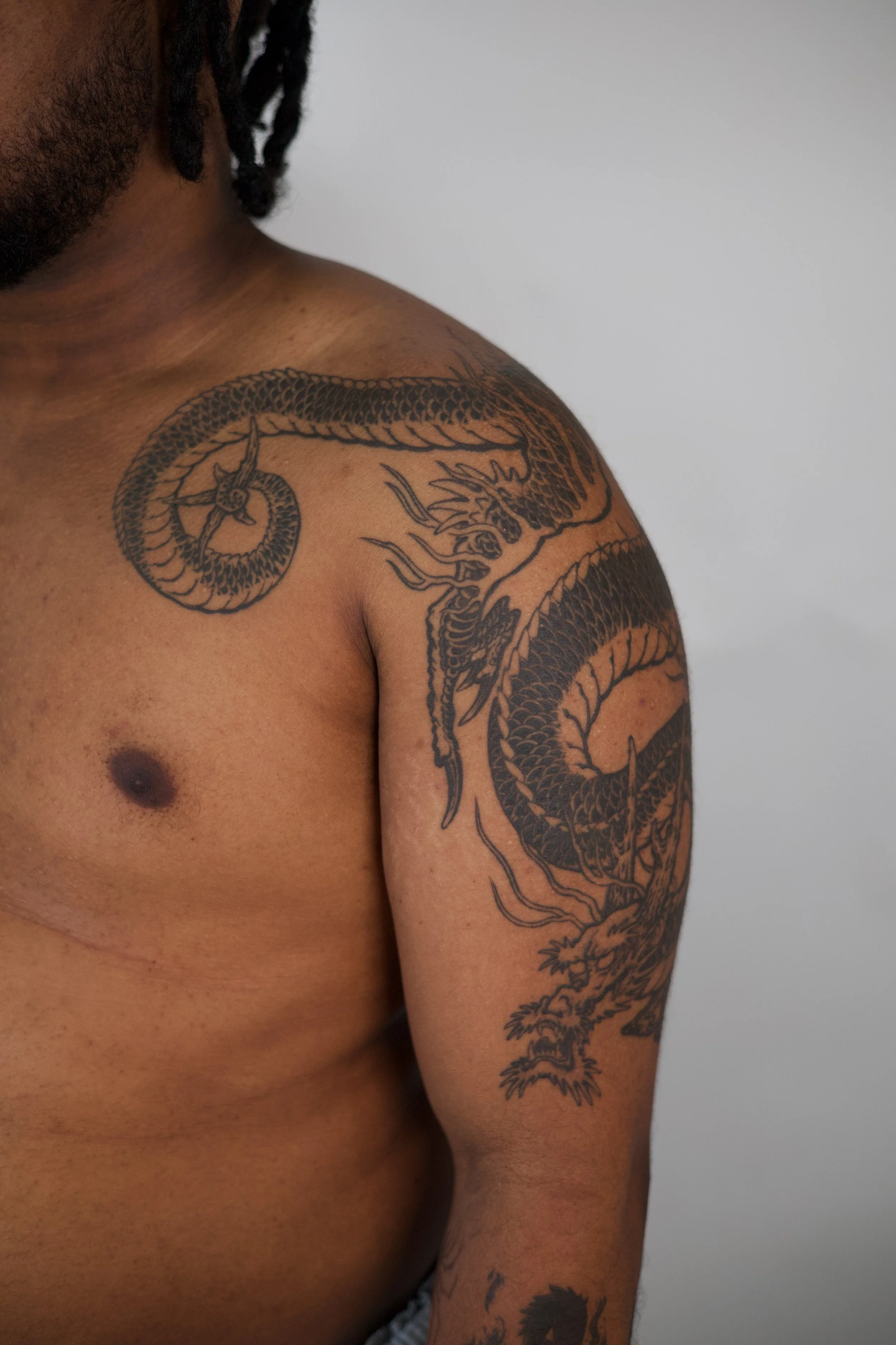 A close-up of a person's upper body showing an elaborate black tattoo of a dragon on their shoulder and upper arm.