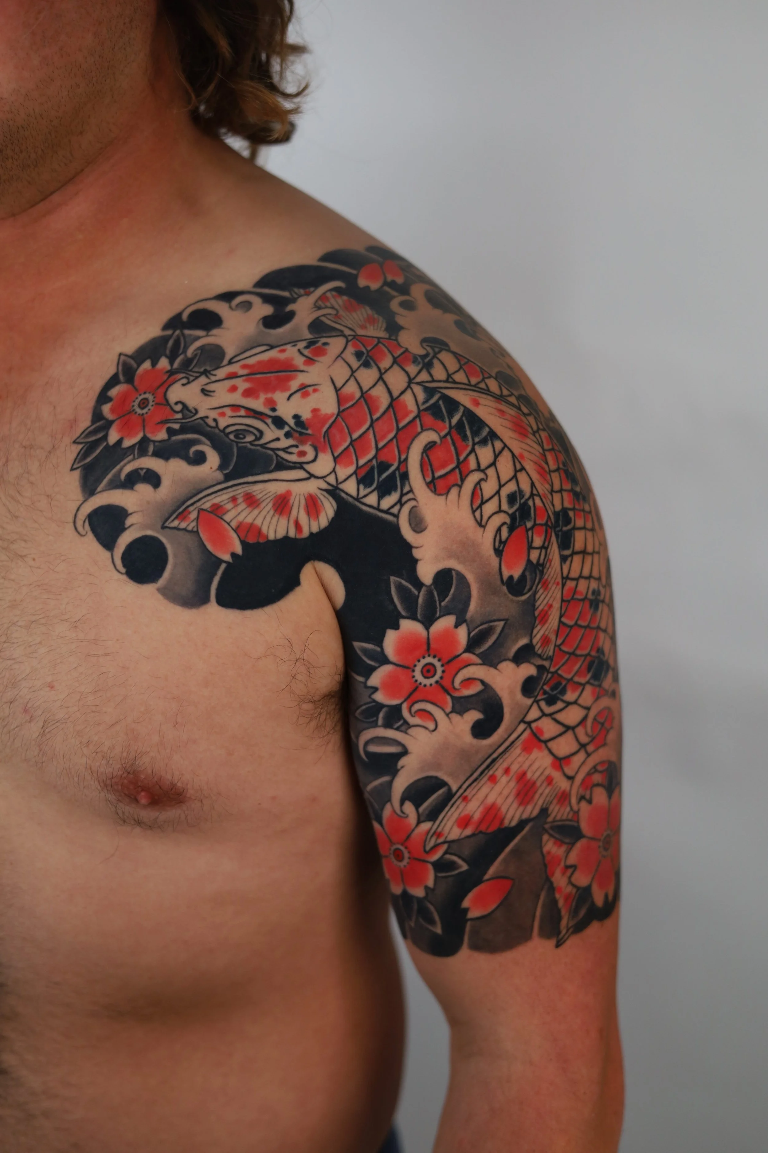 A close-up of a man's upper body and right shoulder displaying a Japanese-style tattoo sleeve featuring a koi fish, flowers, and traditional cloud and wave patterns in black, red, and beige ink.