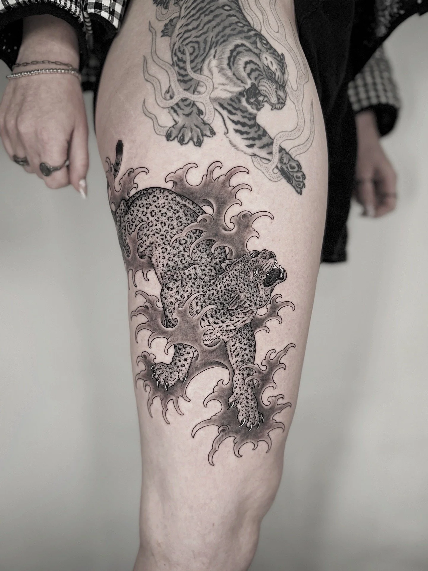 Tattoo of a fierce tiger above a jumping leopard surrounded by stylized waves on a person's thigh.