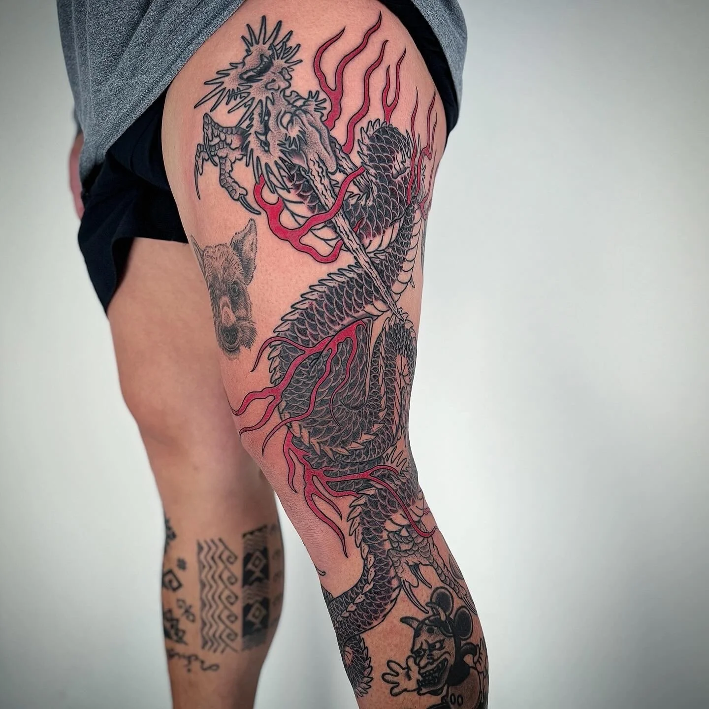 A person's leg with multiple tattoos, including a large black and red dragon wrapping around the thigh and calf, a small bear face on the upper thigh, and a Mickey Mouse tattoo on the lower leg.