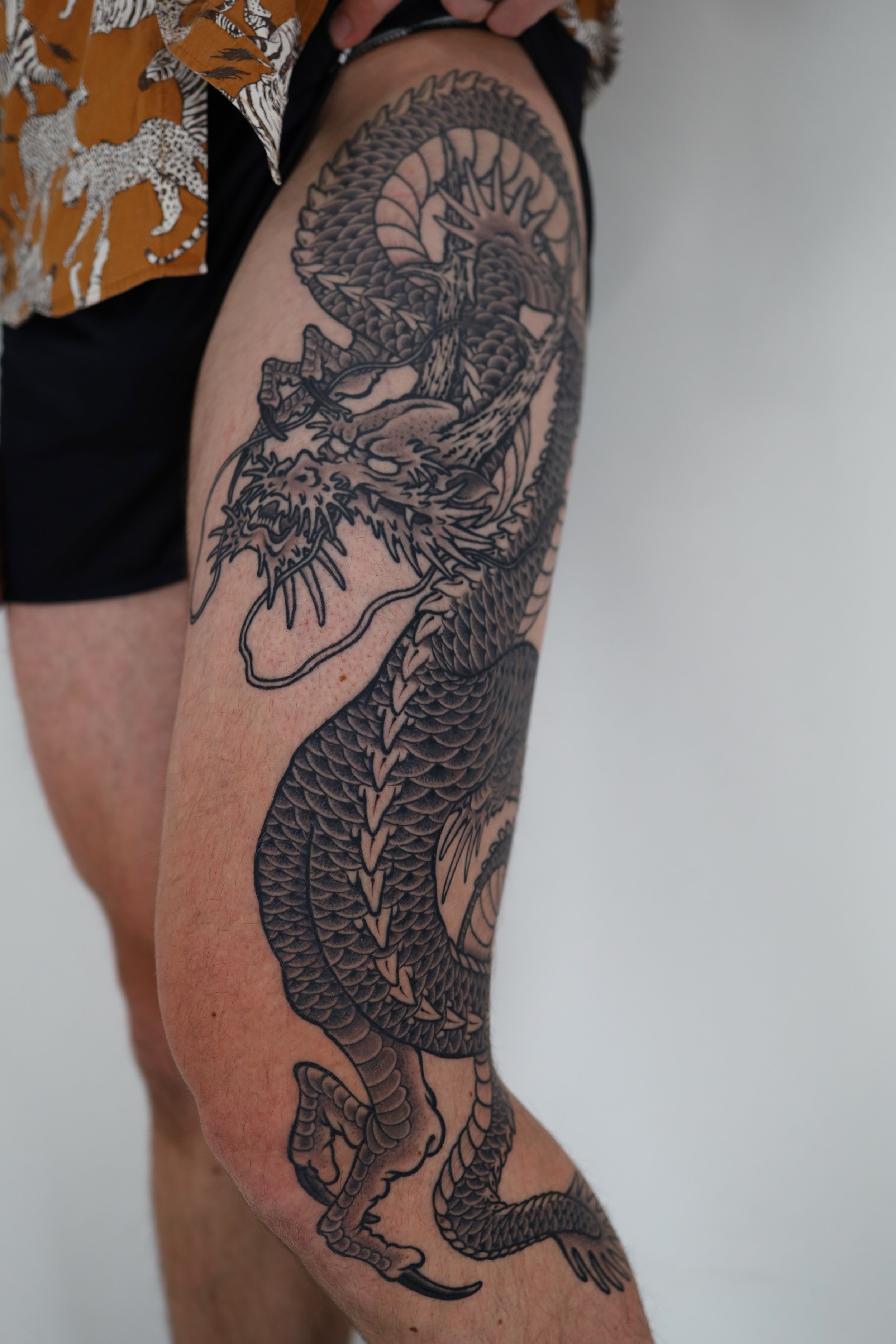 Black outline tattoo of a fierce dragon on a person's arm, with detailed scales and sharp claws, extending from the shoulder to the forearm.