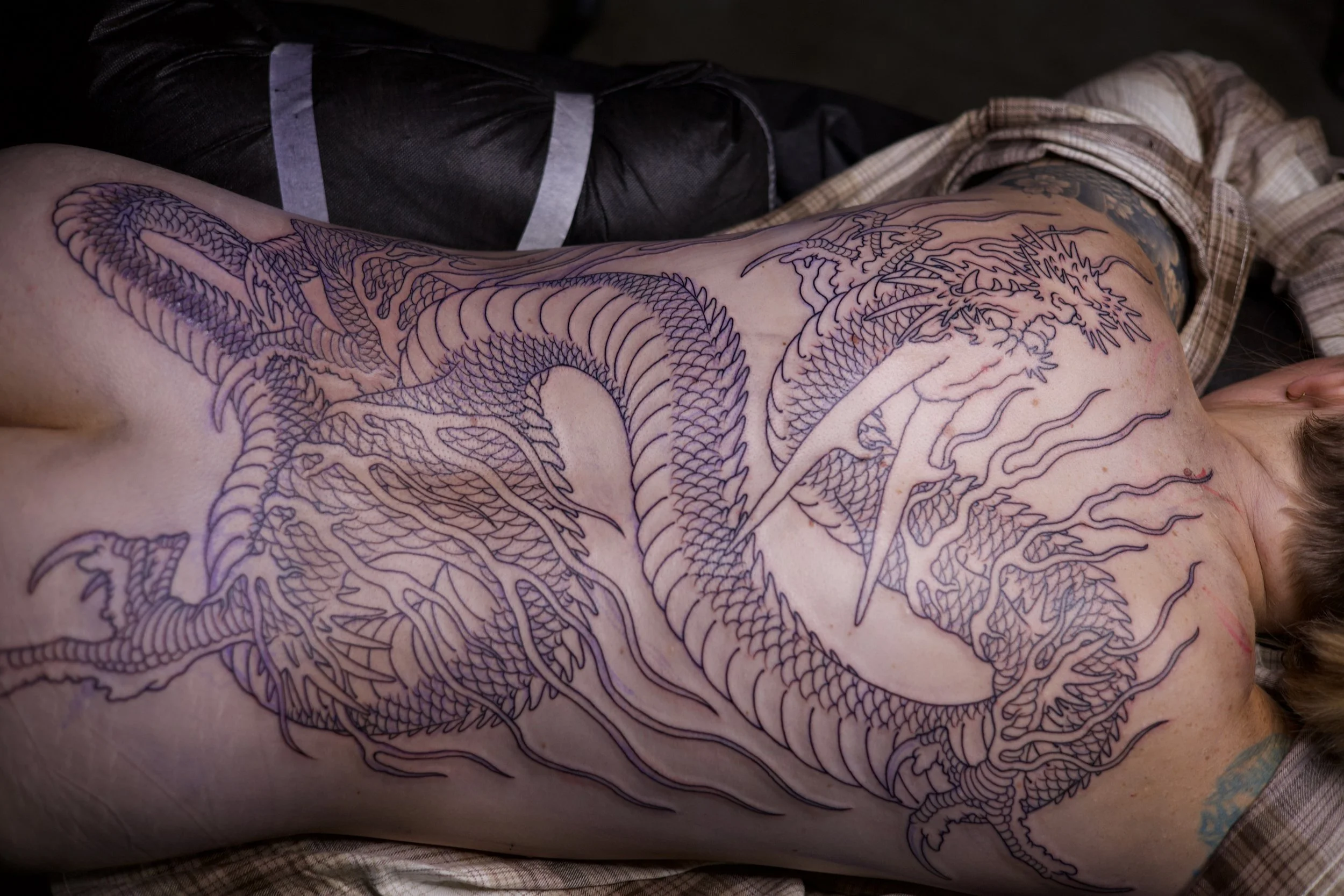 A person lying on their side with a large dragon tattoo outlined on their back, chest, and shoulder.