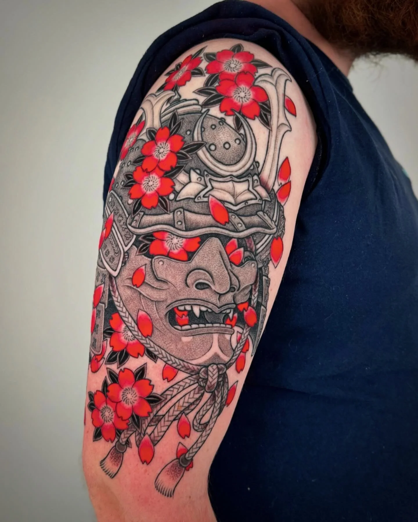 Tattoo of a Japanese demon mask with gray shading and red flowers with leaves, on person's upper arm.