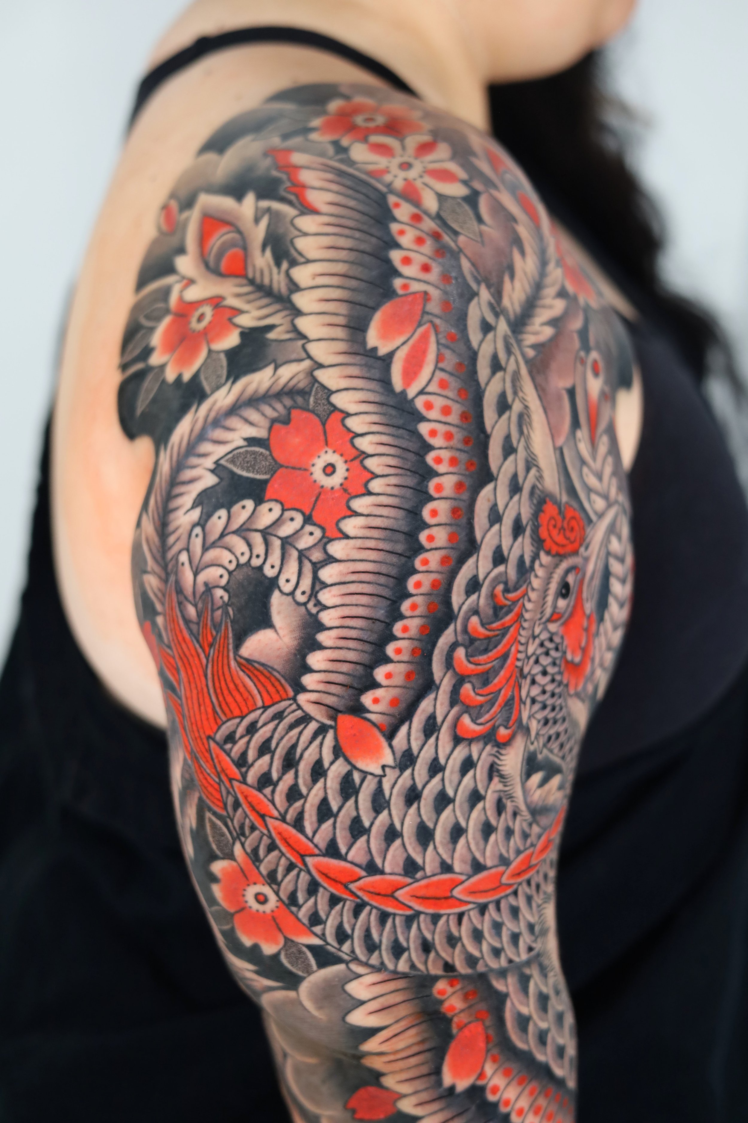 Close-up of a person's shoulder and upper arm tattoo featuring a dragon in black, gray, and red-orange colors with floral and leaf motifs.