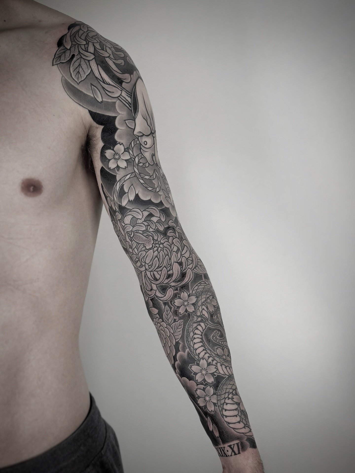 A black and gray full sleeve tattoo featuring flowers, a snake, and a face on a person's arm.