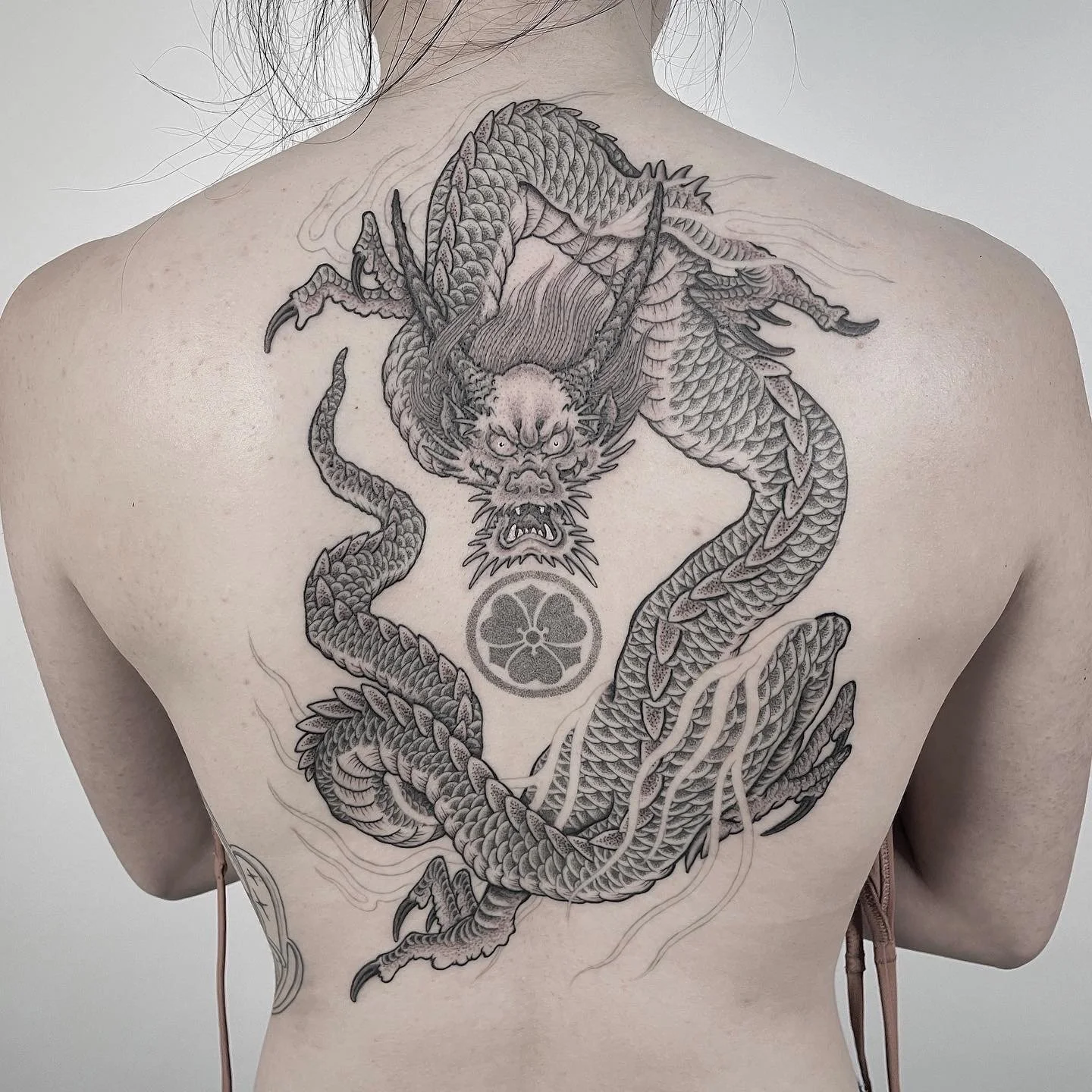 A detailed black and gray tattoo of a fierce dragon on a person's back, with the dragon's body coiled around a circular symbol.