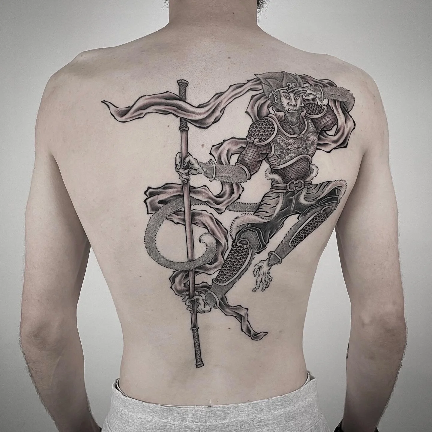 A large black and gray tattoo of a mythological warrior with a lion's face, dressed in armor and holding a staff, is inked on a man's back.