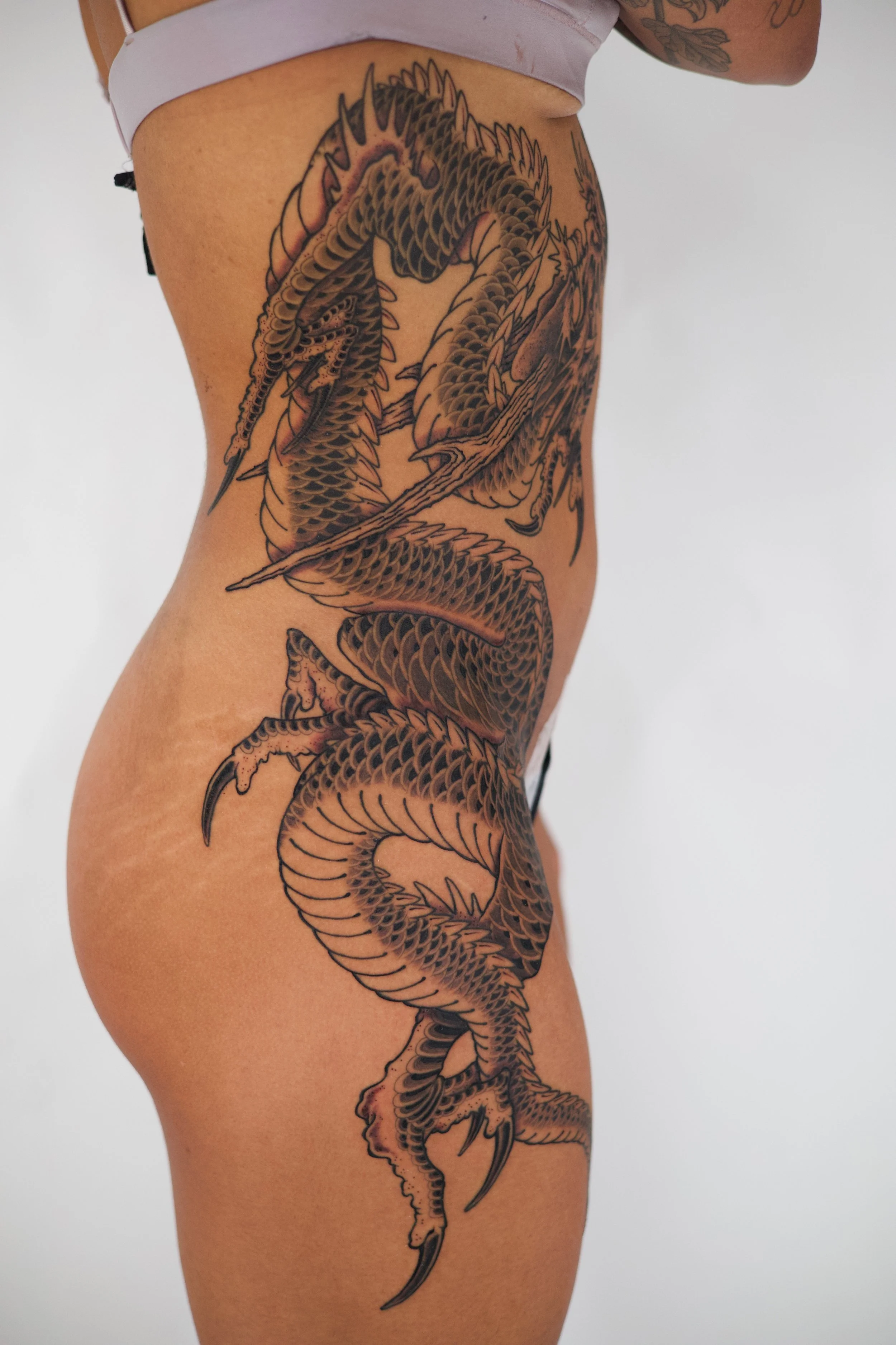 A large, detailed black and gray tattoo of a dragon on a woman's side, extending from her waist to her thigh.