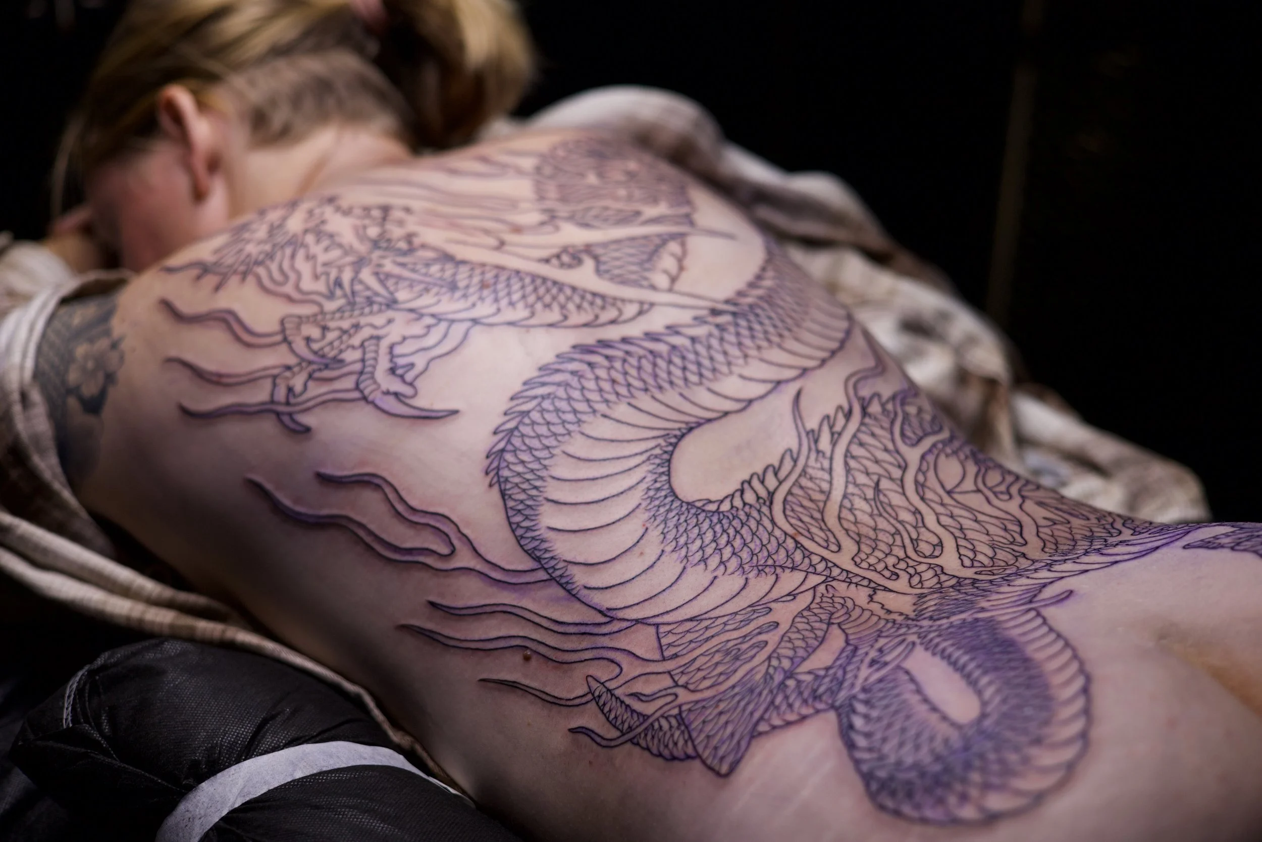 A person lying on an bed with a large dragon tattoo on their back, outlined in purple ink.